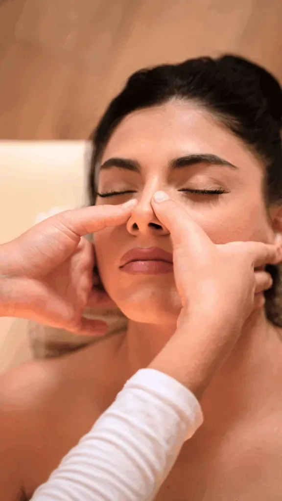 A woman receiving a facial massage on her nose from a person with hands in a clinical or spa setting.