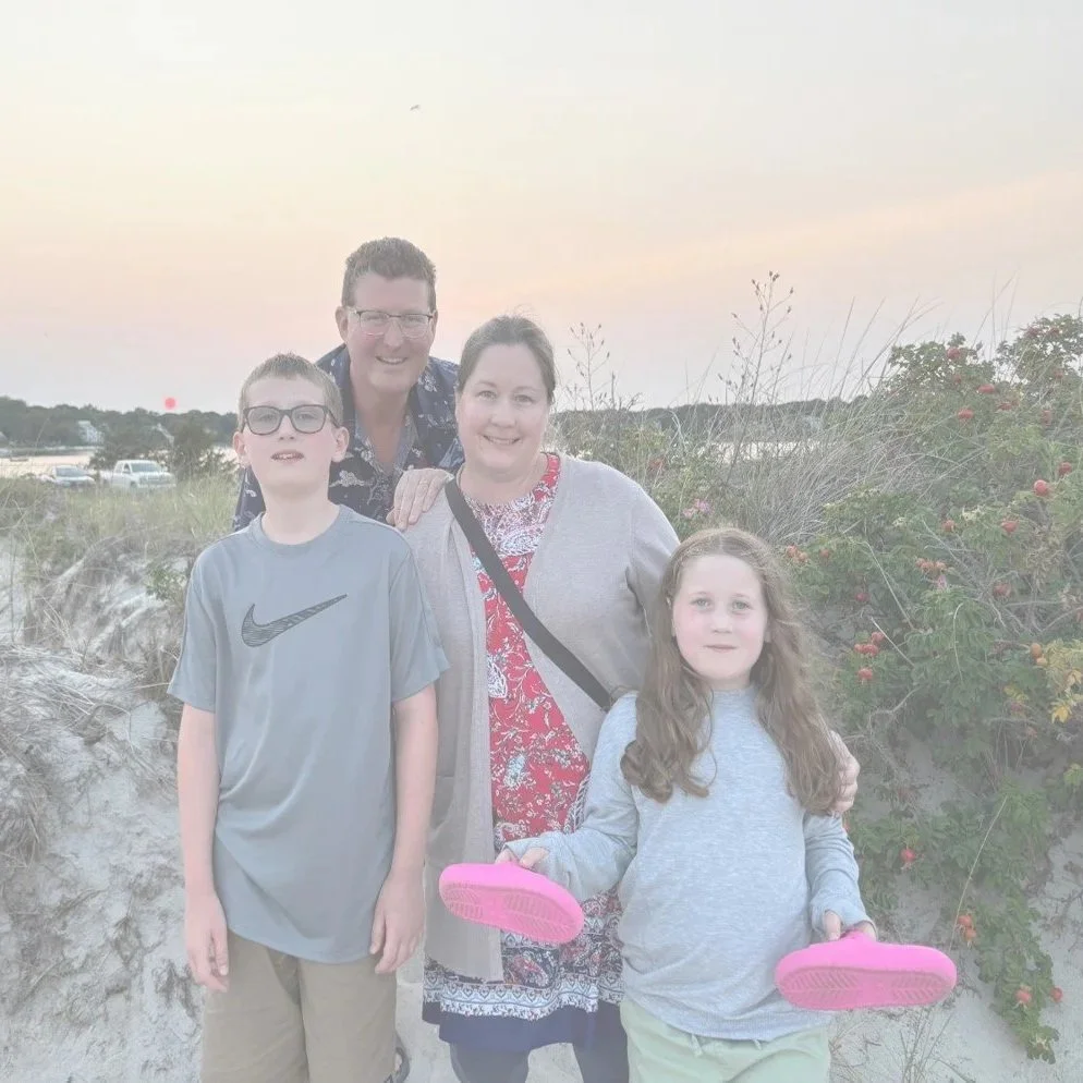 A family of four standing outdoors on a beach at sunset, with a man and woman smiling behind a boy wearing glasses and a girl with long hair holding pink flip-flops.