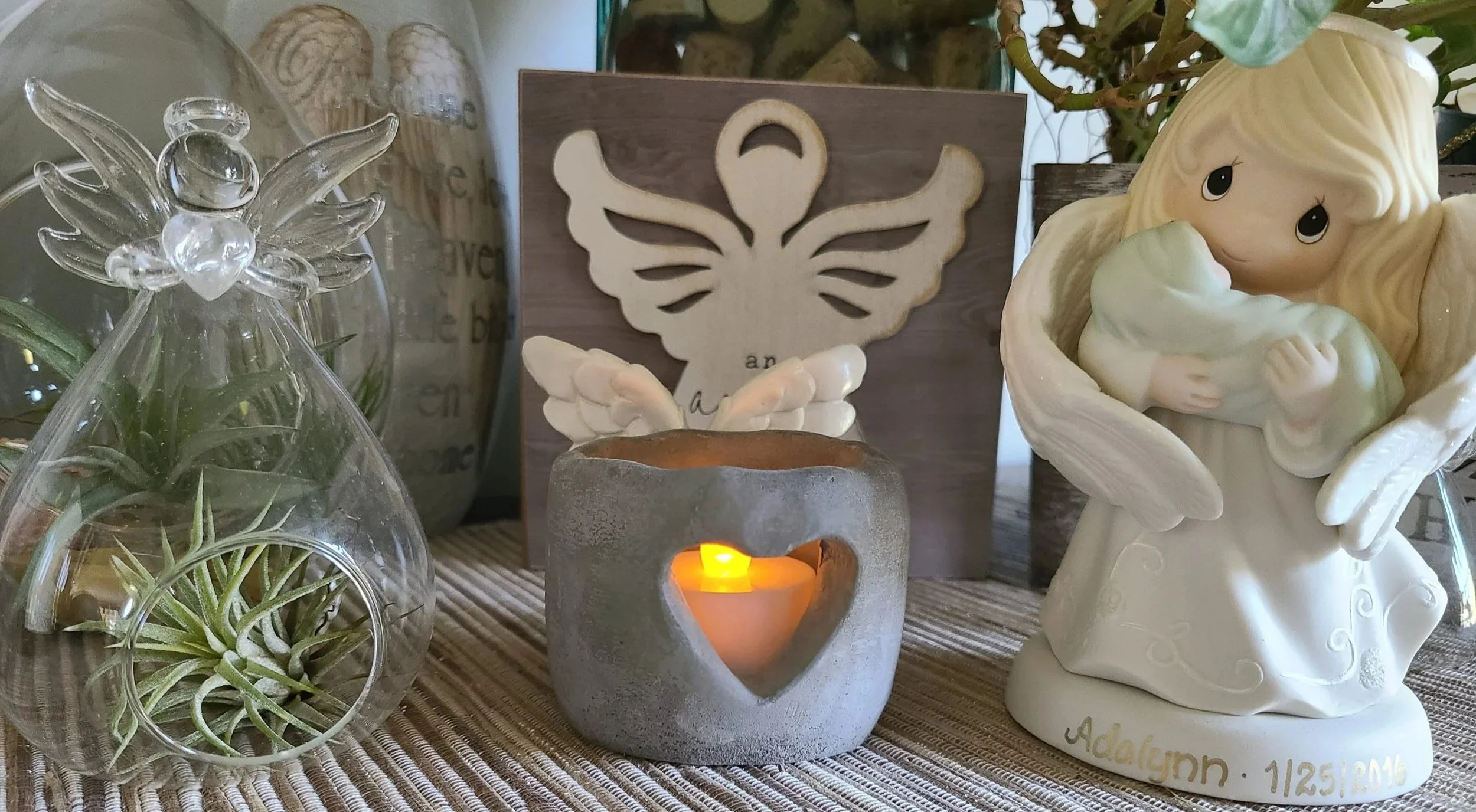 Decorative arrangement on a table featuring a small glass angel figurine, a candle holder with a lit candle, a wooden cutout of a butterfly with angel wings, a small potted plant, and a ceramic angel figurine holding a flower. The scene is set on a woven table runner.