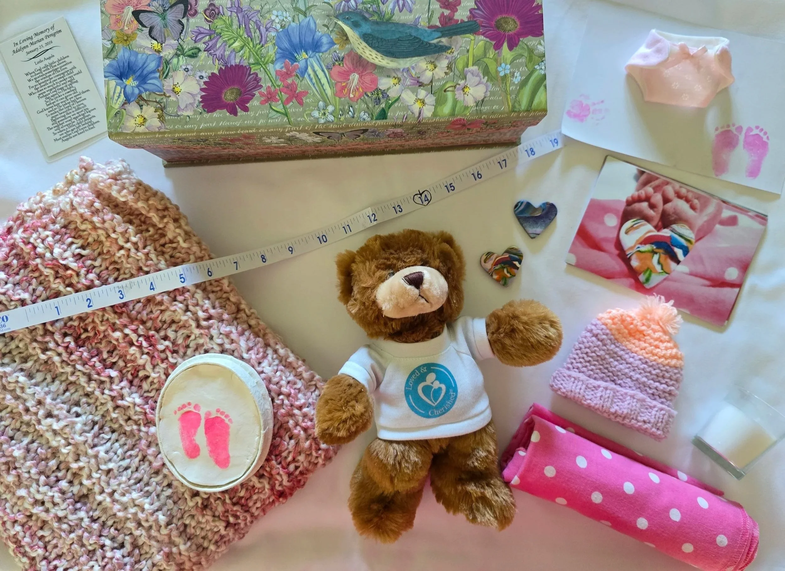 A collection of items including a handmade pink and white crochet blanket, a teddy bear wearing a white shirt with a blue circular logo, pink footprints on a paper, a pink and white polka dot sock, a small pink sock with a frilly top, a mirror with baby footprints, a plush star, a pink card with footprints, a floral jewelry box, a small heart-shaped object, a ruler, a paper with a poem or prayer, and a floral-illustrated box with a bird on it.