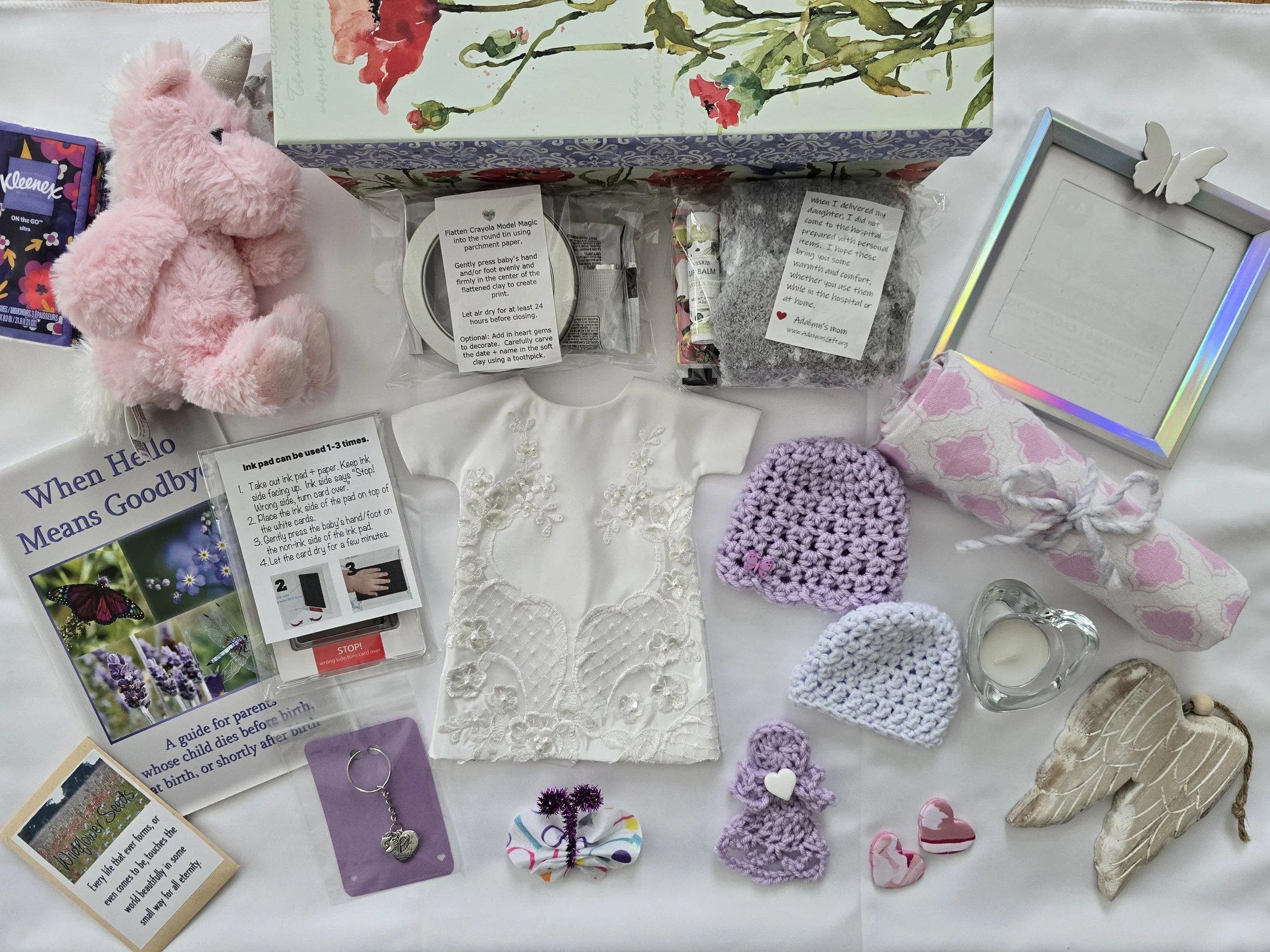 Collection of baby items on a wooden floor, including a plush bunny toy, a baby dress, hats, crocheted hearts, a blanket, books, and personal care products.