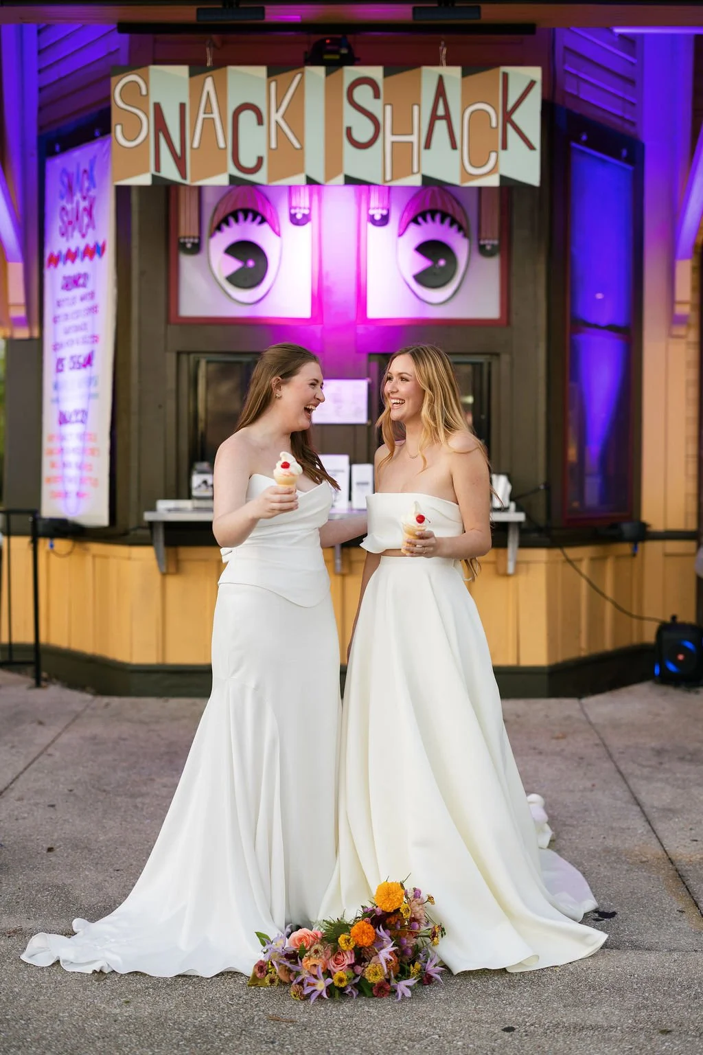 Two brides eating soft-serve ice cream cones outside a window concession stand. The building reads "Snack Shack"