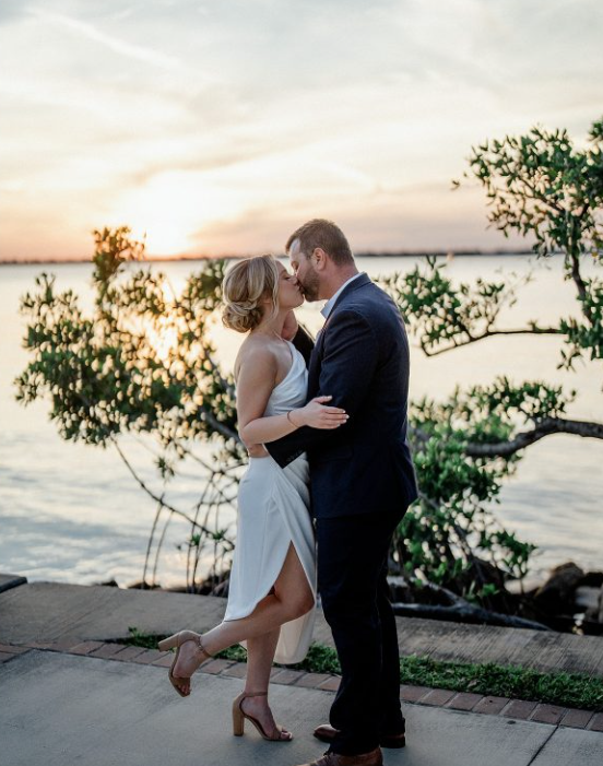 A couple in wedding attire sharing a kiss by the water at sunset, with trees in the background.