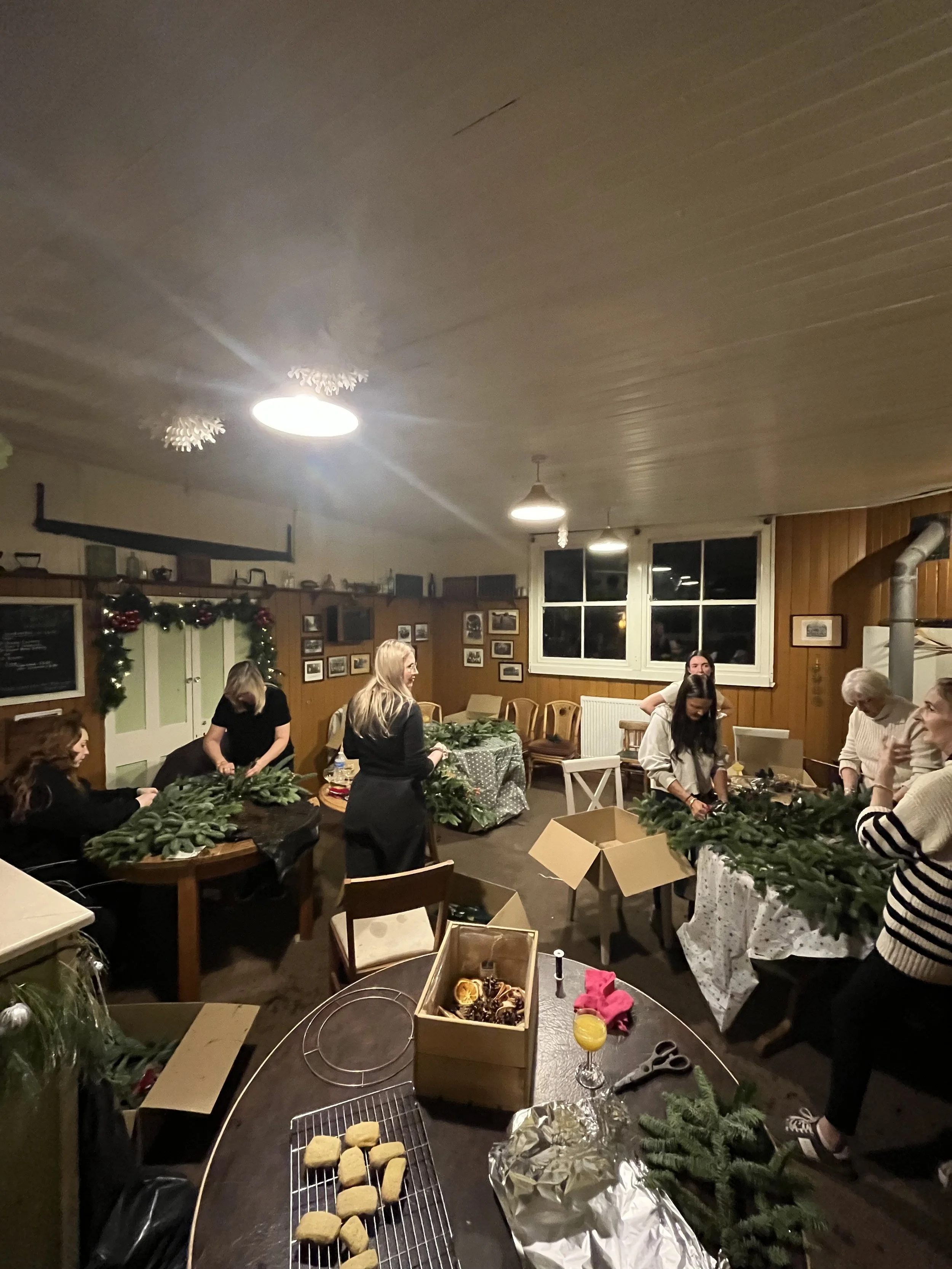People gathered around a table preparing Christmas cookies and decorating with evergreen branches in a warmly lit room.