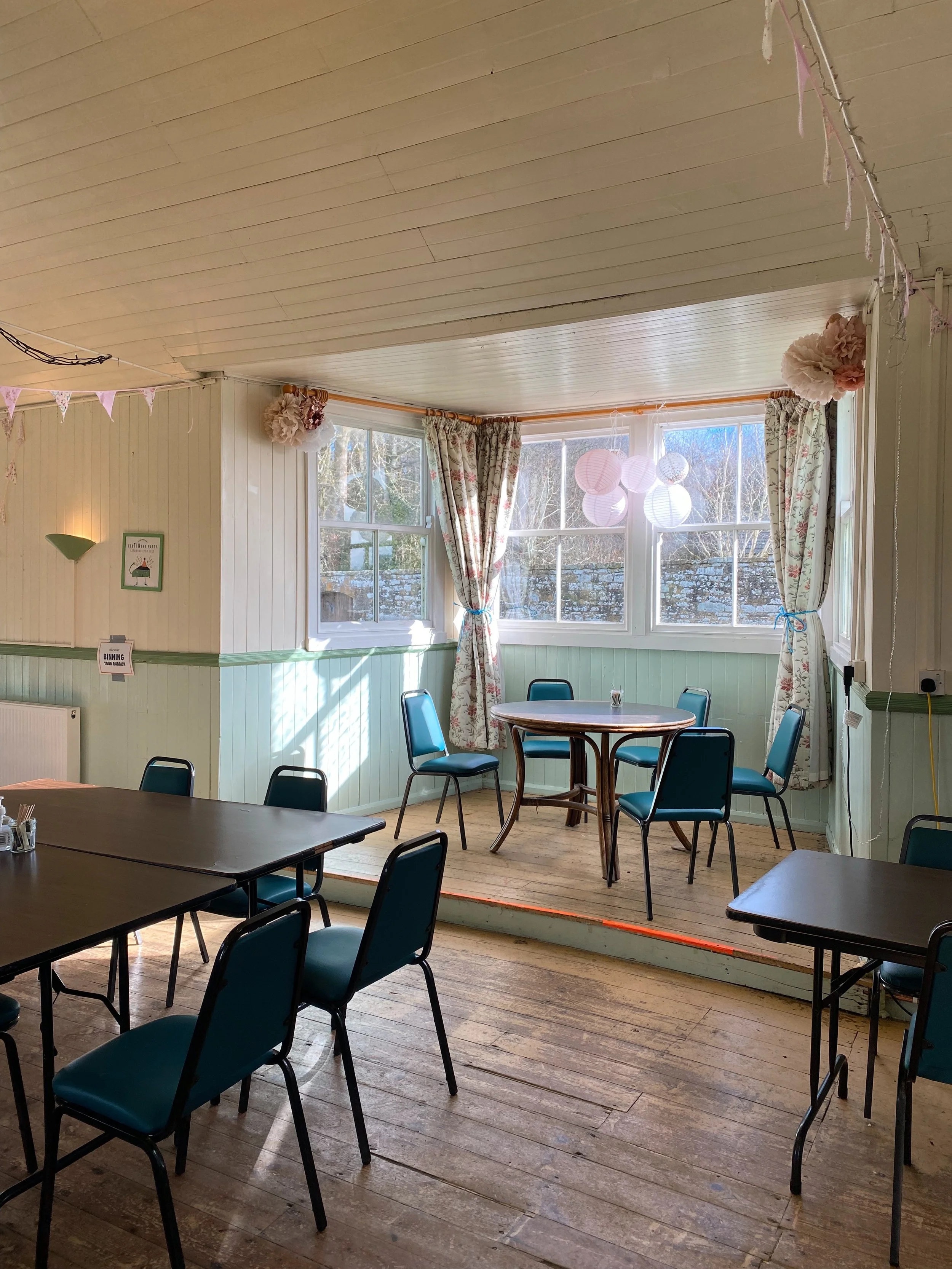A cozy dining area with large windows, floral curtains, and pastel decorations including paper lanterns and tissue paper pom-poms, with several tables and chairs for seating.