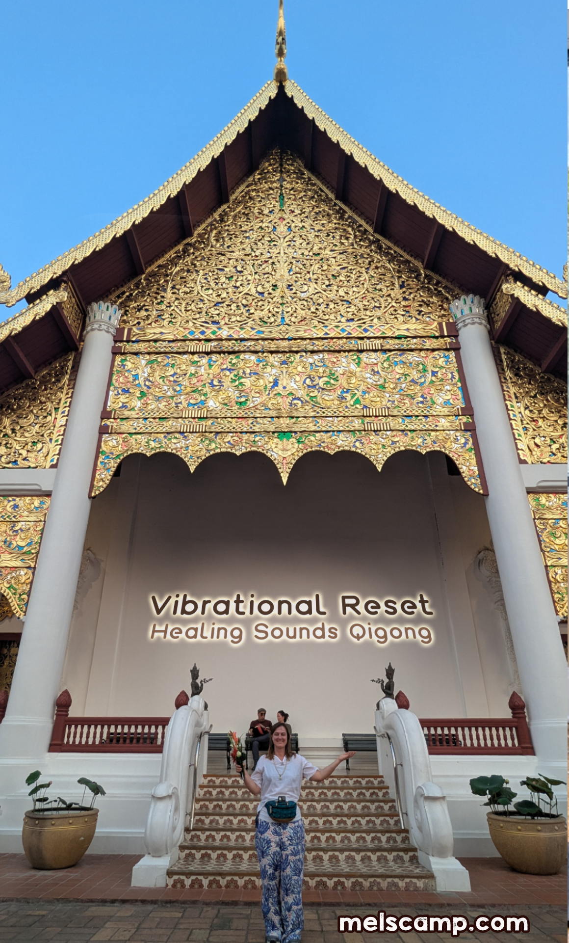 A woman standing on stairs in front of a temple entrance in Chiang Mai Thailand on Mahabuka Day with ornate gold decoration, holding a flower and smiling. The temple has a sign that reads "Vibrational Reset Healing Sounds Qigong."