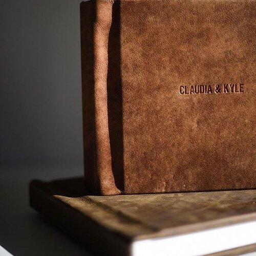 Close-up of a suede-covered book with the text 'Claudia & Kyle' embossed on the cover, resting on another book.
