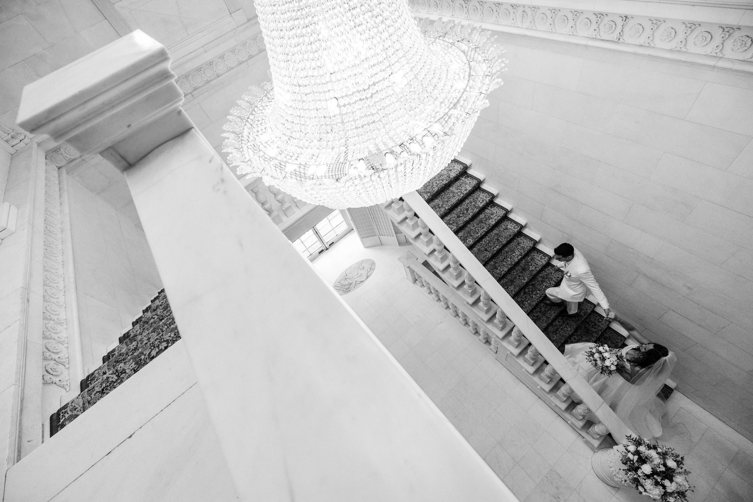 Black and white aerial view of a bride and groom descending a staircase in an elegant building with a large chandelier overhead.
