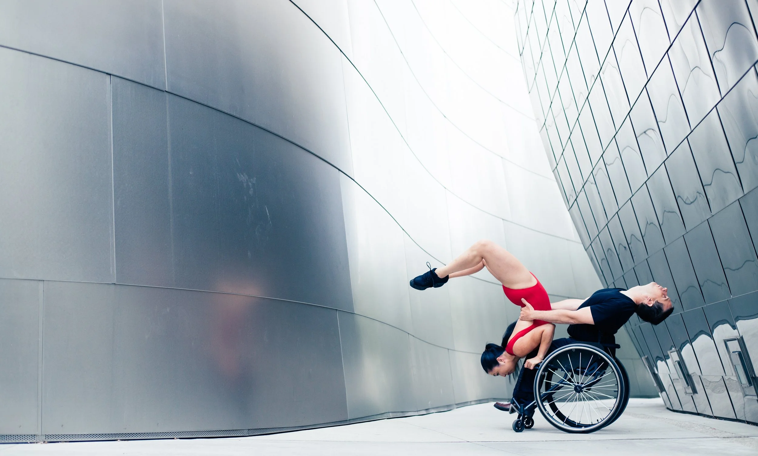 Dance photo. Marisa arches over a male wheelchair dancer's lap as if she is doing a handstand scorpion