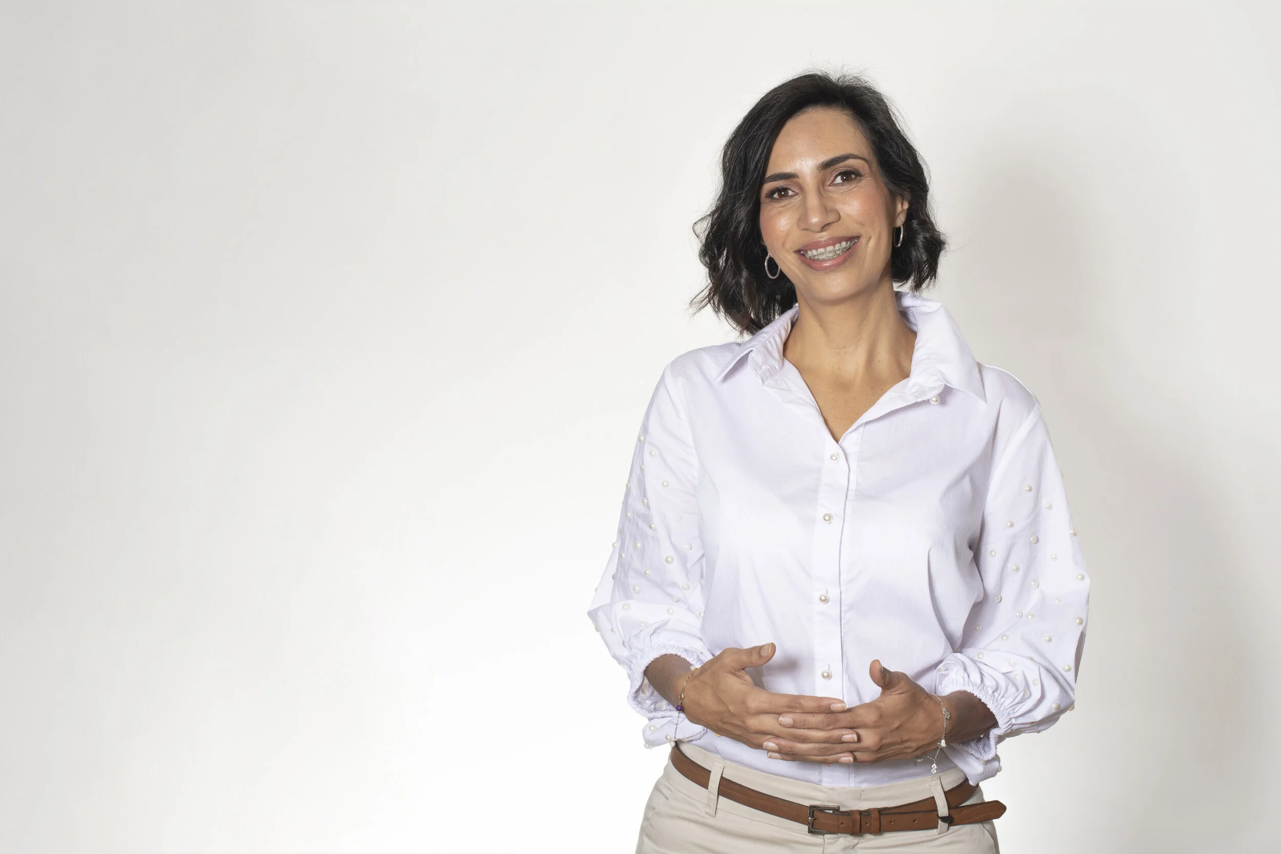 Mujer con cabello oscuro, sonriendo y usando blusa blanca con detalles en perlas, de fondo blanco.