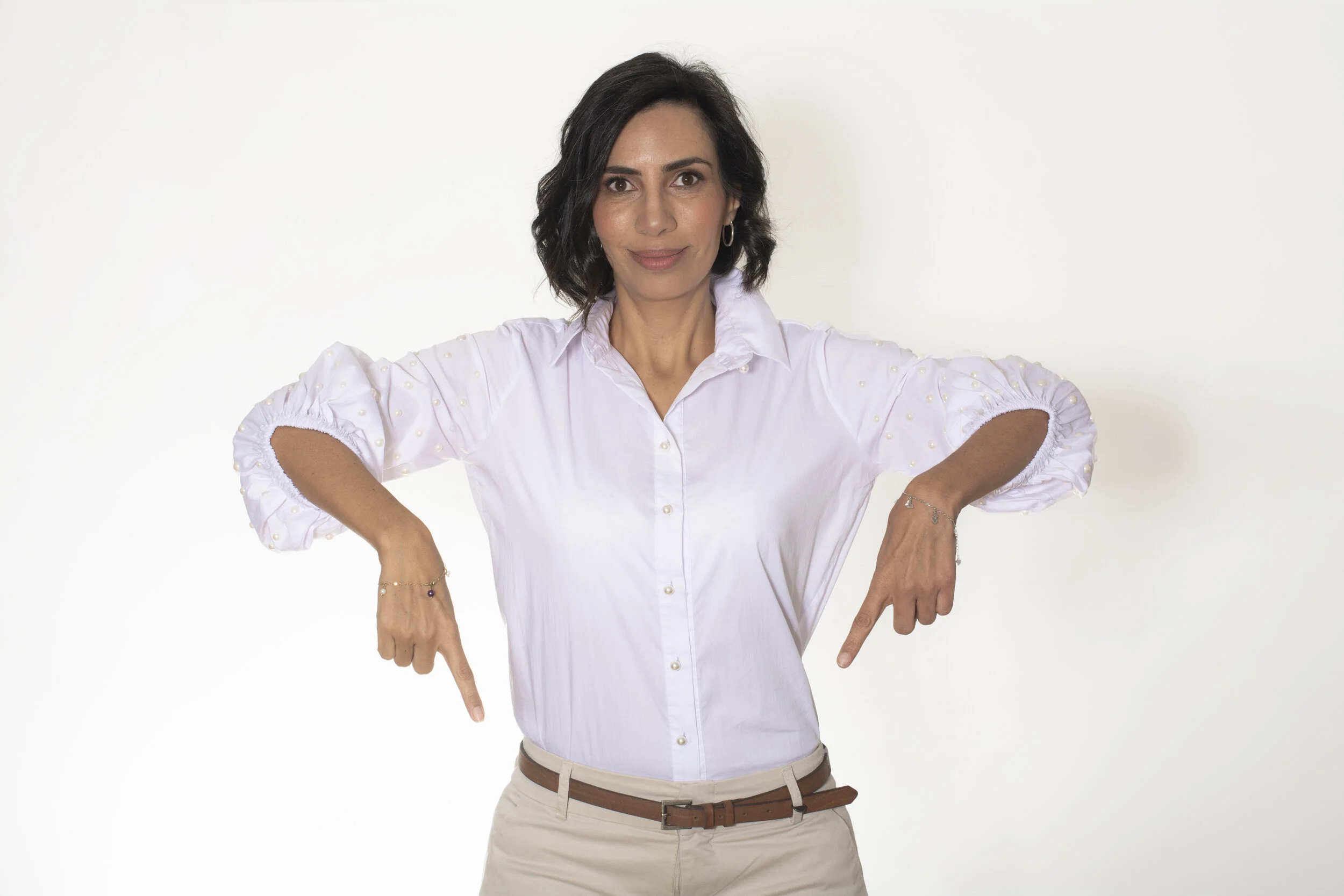 Mujer con cabello oscuro, camiseta blanca con detalles de perlas y pantalones beige, señalando hacia abajo con ambas manos, en un fondo blanco.