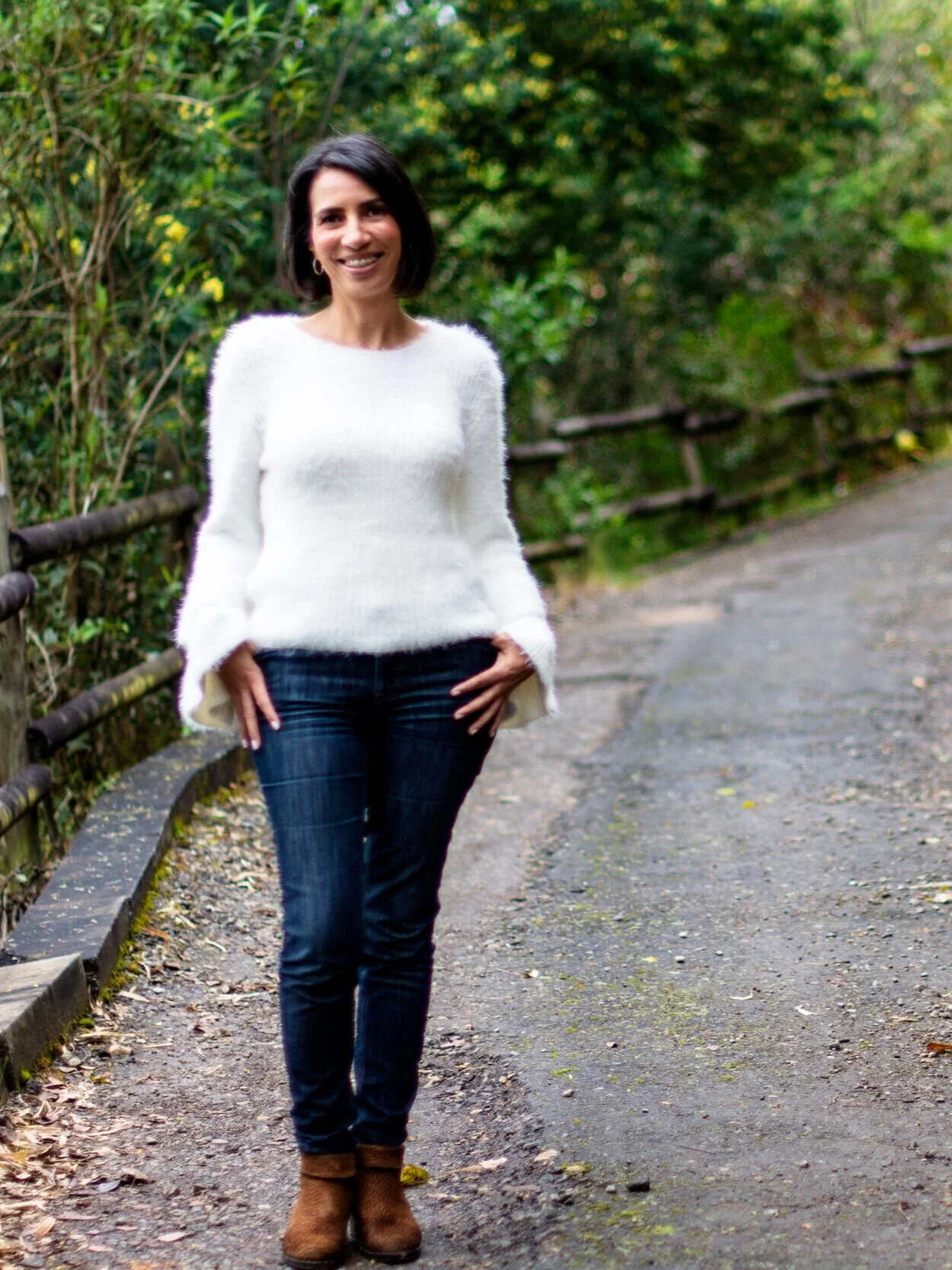 Mujer con cabello corto, camiseta blanca y pantalones azules, caminando por un sendero rodeado de árboles verdes.