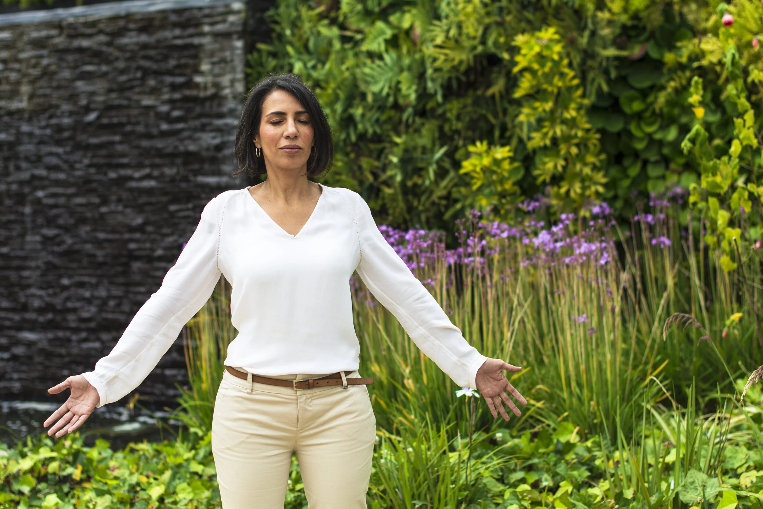 Mujer con los ojos cerrados en postura de meditación en un jardín con plantas verdes y flores moradas.