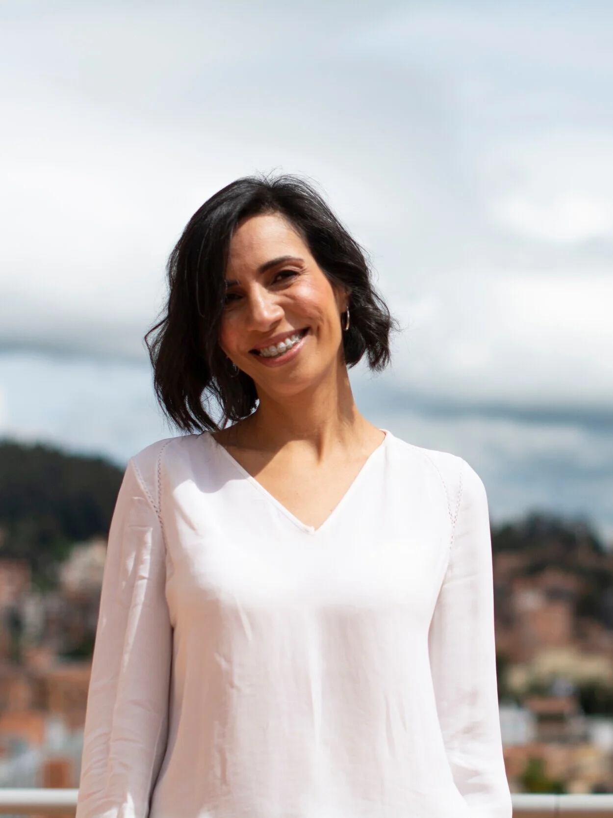 Mujer con cabello castaño oscuro corto, sonriendo, lleva blusa blanca, fondo de cielo nublado y vista de ciudad.