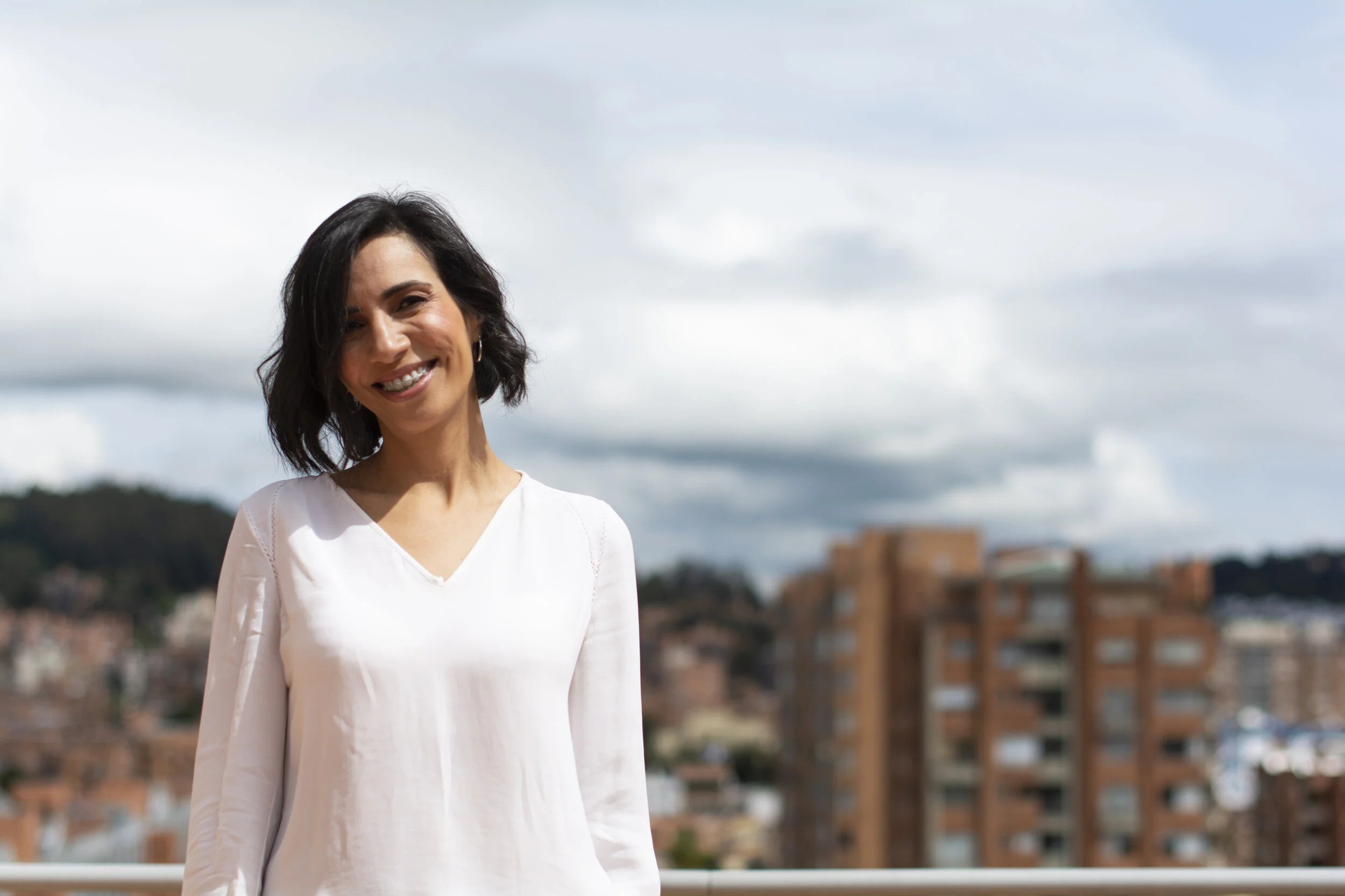 Mujer sonriente con cabello corto y oscuro, vistiendo una blusa blanca, de pie en un espacio con fondo de cielo nublado y edificios urbanos.