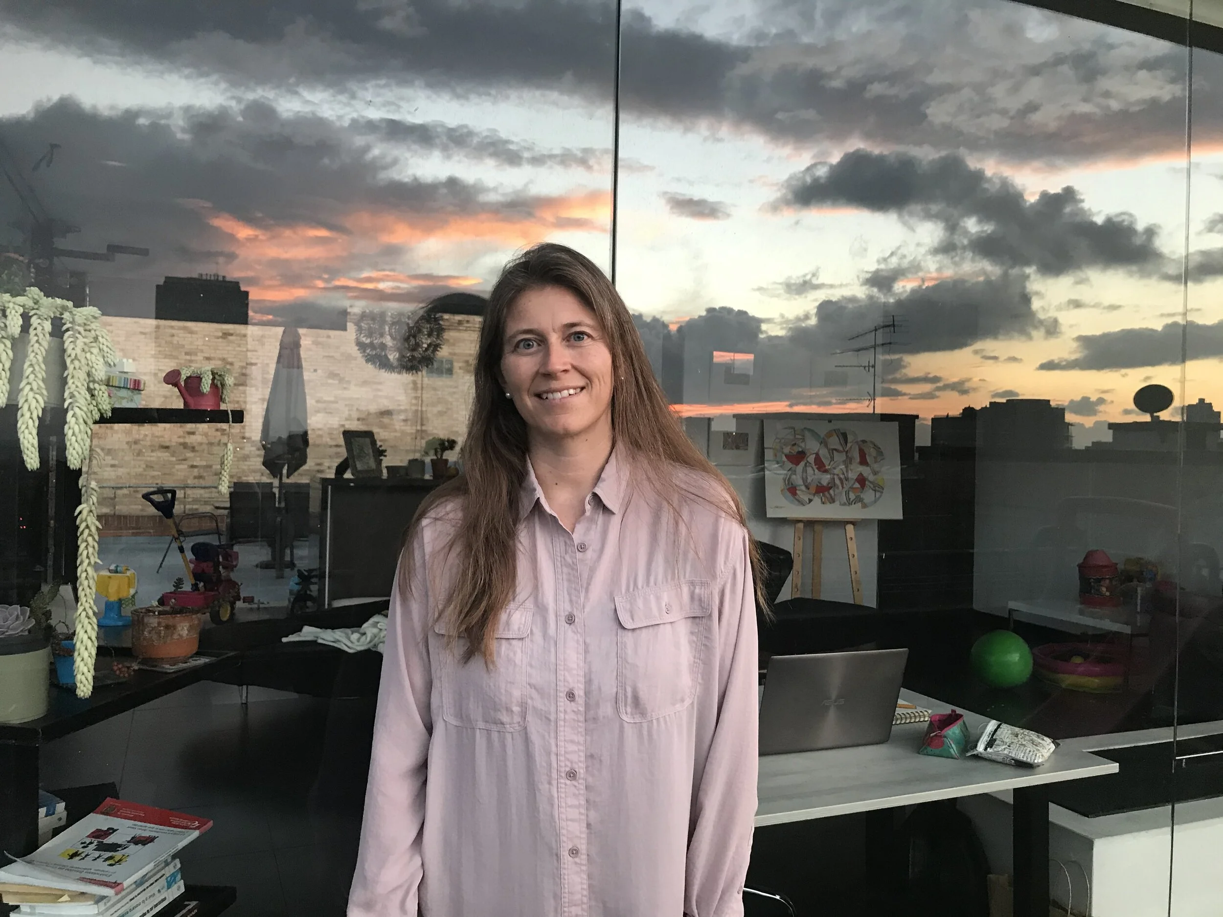 Una mujer con cabello largo y rubio claro, vestida con una camisa de botones color rosa claro, de pie en un espacio interior con una pared de vidrio que refleja un cielo nublado con tonos rosados y anaranjados en el atardecer.