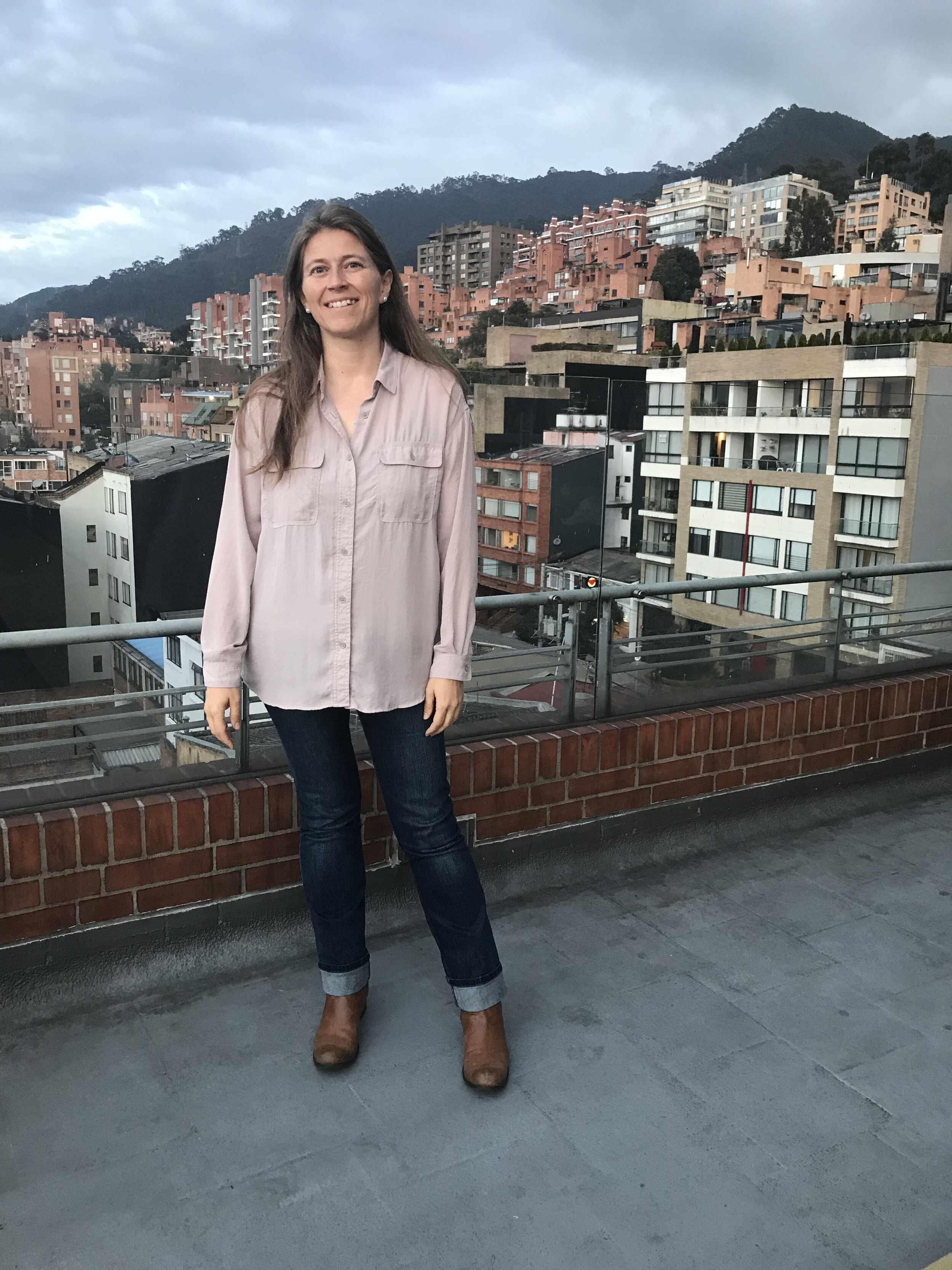 Mujer sonriendo con paisaje de edificios y montañas en el fondo en un balcón.