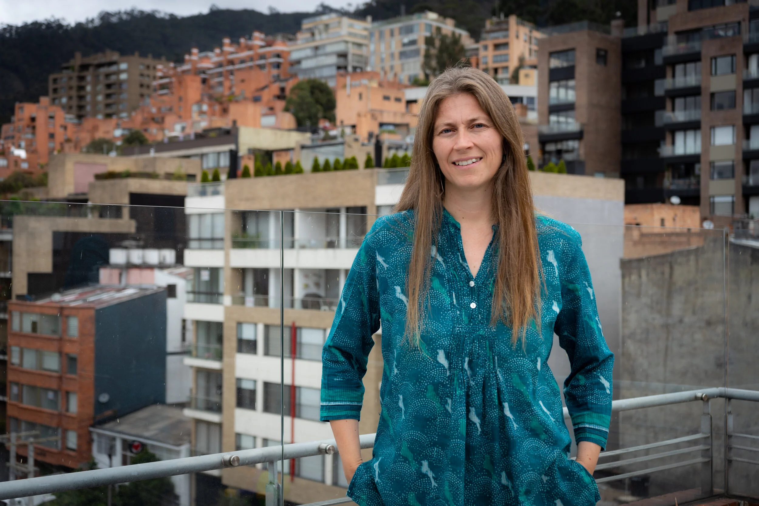 Mujer con cabello largo y liso, sonriendo, de camiseta azul con estampado, en una terraza con vista a edificios y colinas en el fondo.