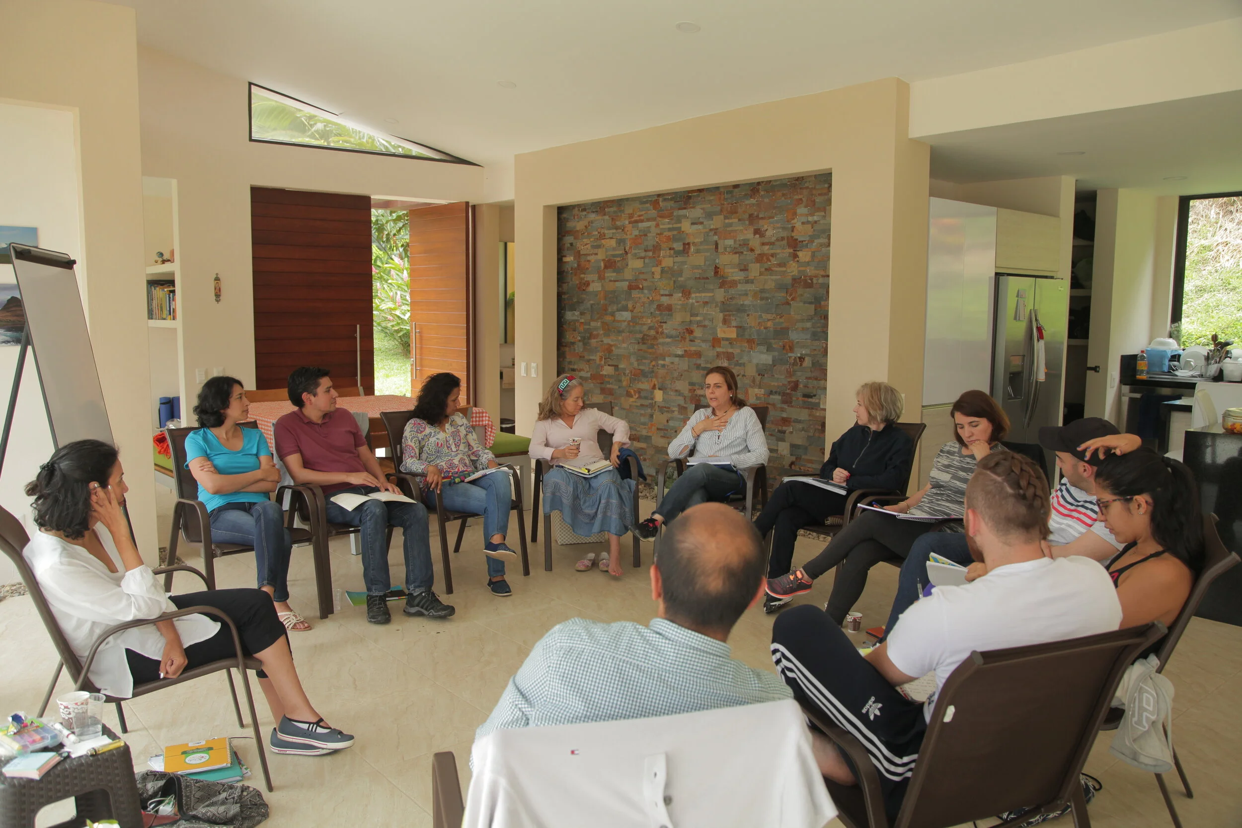 Grupo de personas sentadas en círculo en una sala, participando en una reunión o discusión, con una pared de ladrillos decorativos y una cocina visible al fondo.