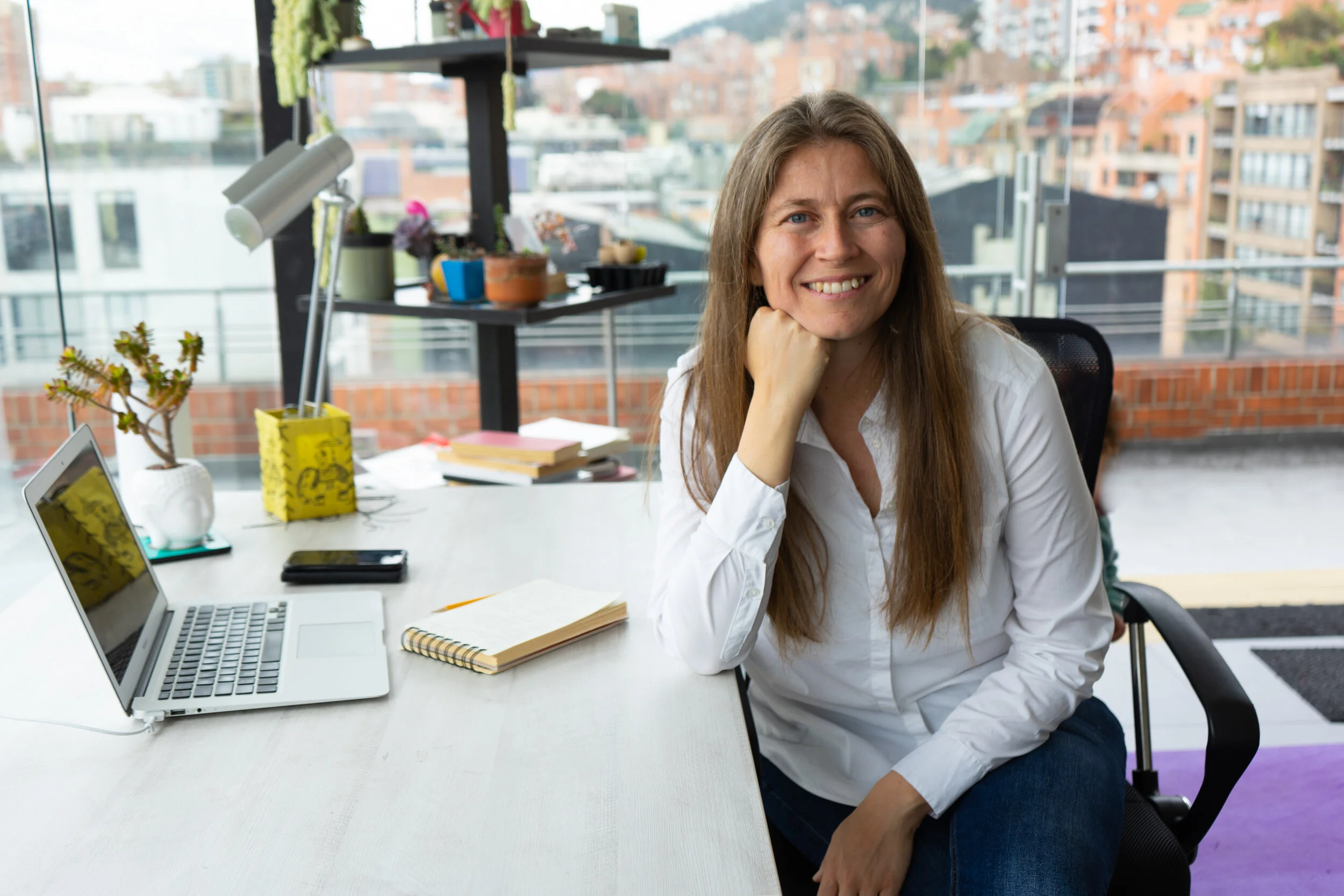 Mujer sonriendo en una oficina moderna con vista a la ciudad, rodeada de una laptop, libros y plantas.