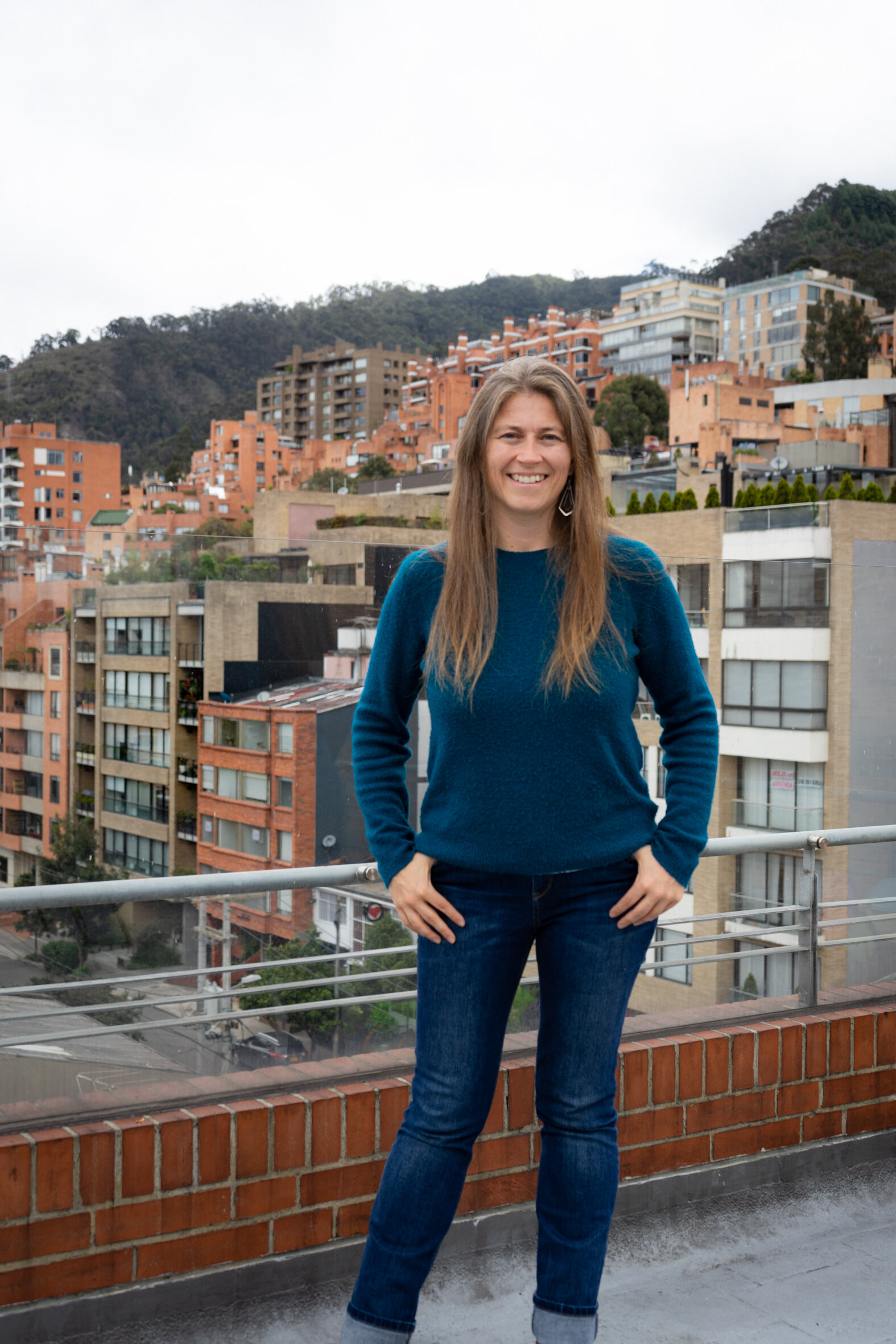 Mujer con cabello largo y rubio, usando un suéter azul y jeans, posando en una terraza con vista a una ciudad en una colina, con edificios color ladrillo y árboles en el fondo.
