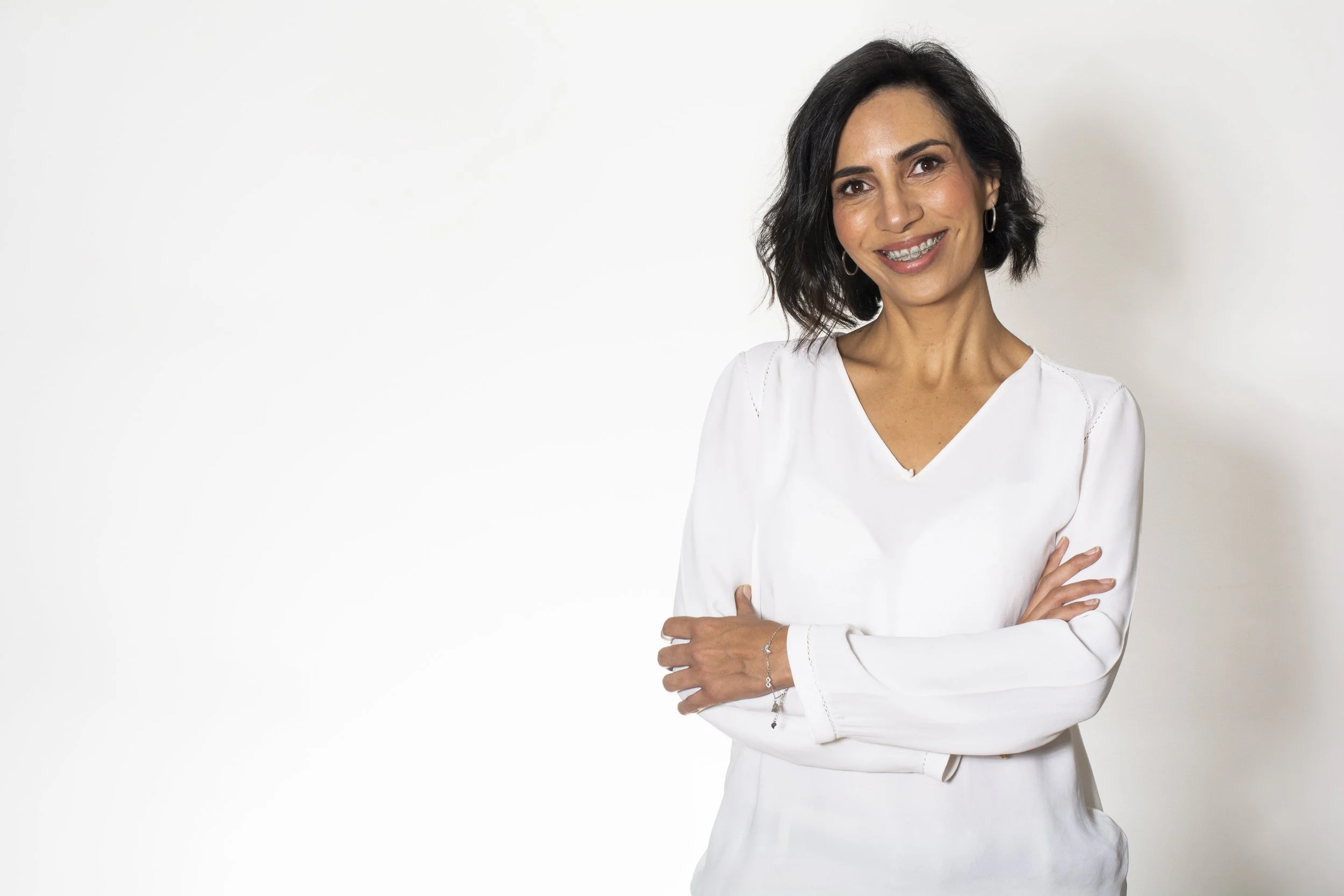 Mujer sonriente con cabello castaño oscuro, usando blusa blanca y accesorios plateados, de pie con brazos cruzados en un fondo blanco.