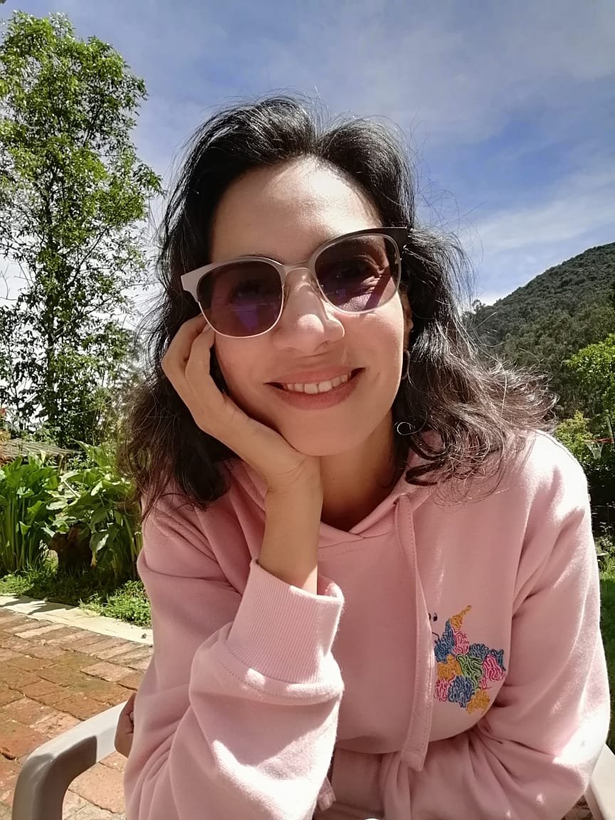Mujer sonriendo con gafas de sol, en un entorno al aire libre con árboles y montañas al fondo, en un día soleado.