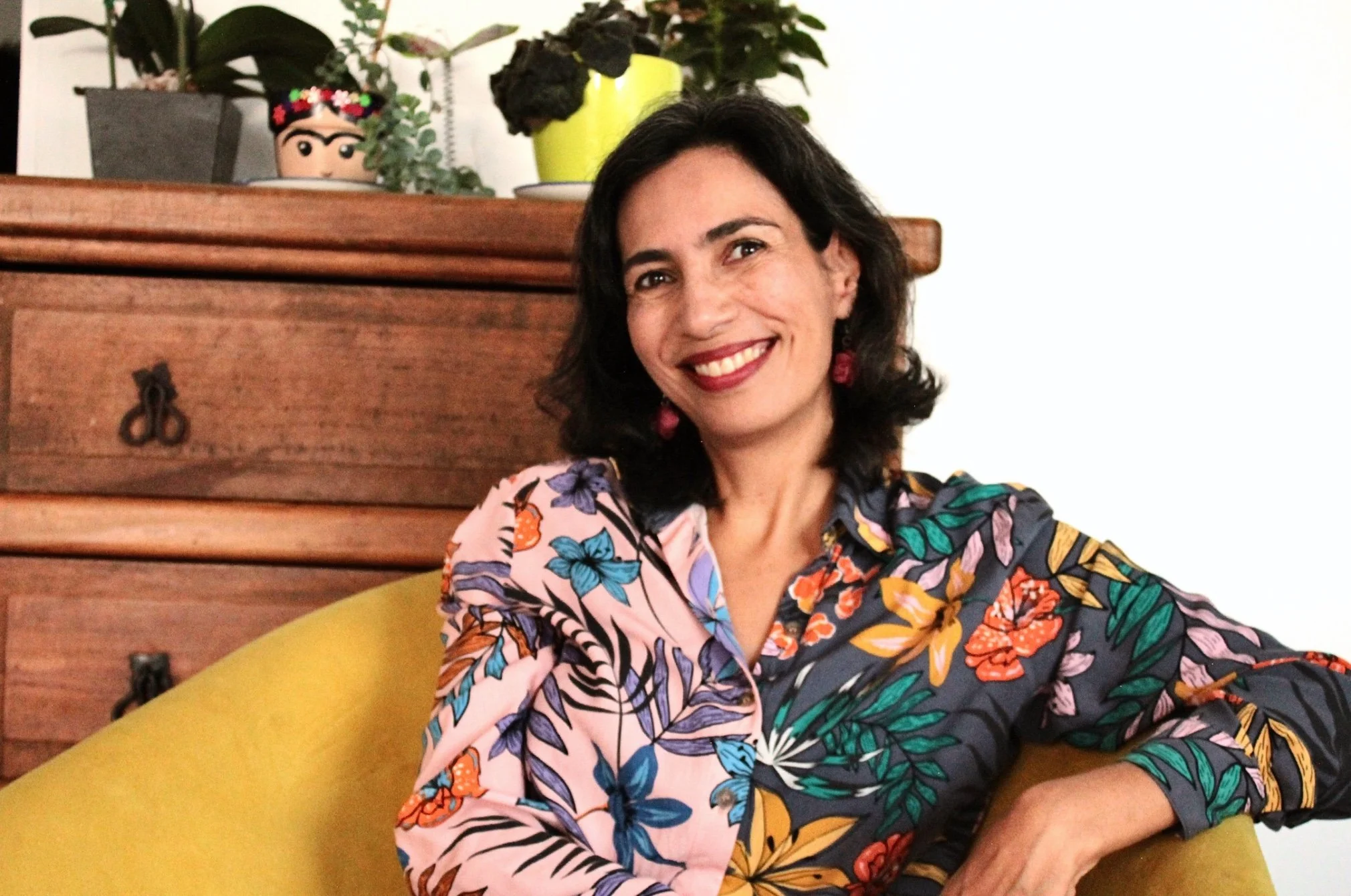Mujer sonriendo con roupa floral sentada en un sillón amarillo, fondo de mesa de madera con plantas decorativas.
