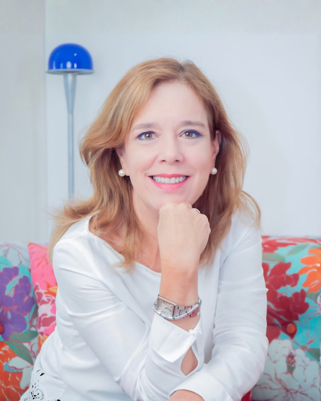 Retrato de una mujer con cabello rubio, sonriendo, usando blusa blanca, con reloj y aretes de perlas, en un entorno con un sofá con cojines coloridos y una lámpara azul.