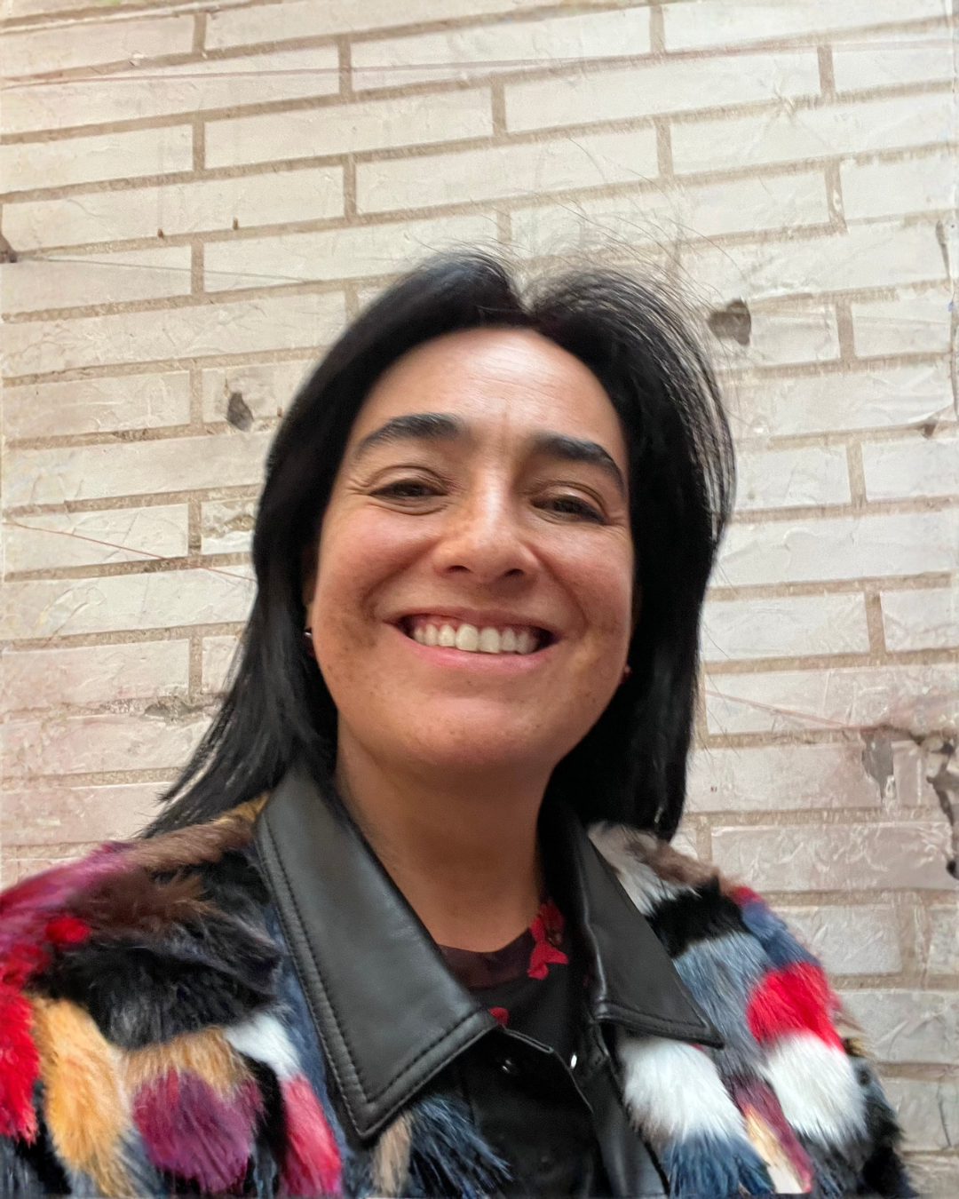 Retrato de una mujer sonriente con cabello negro, usando una chamarra de cuero con detalles de piel en las mangas, frente a una pared de ladrillos.