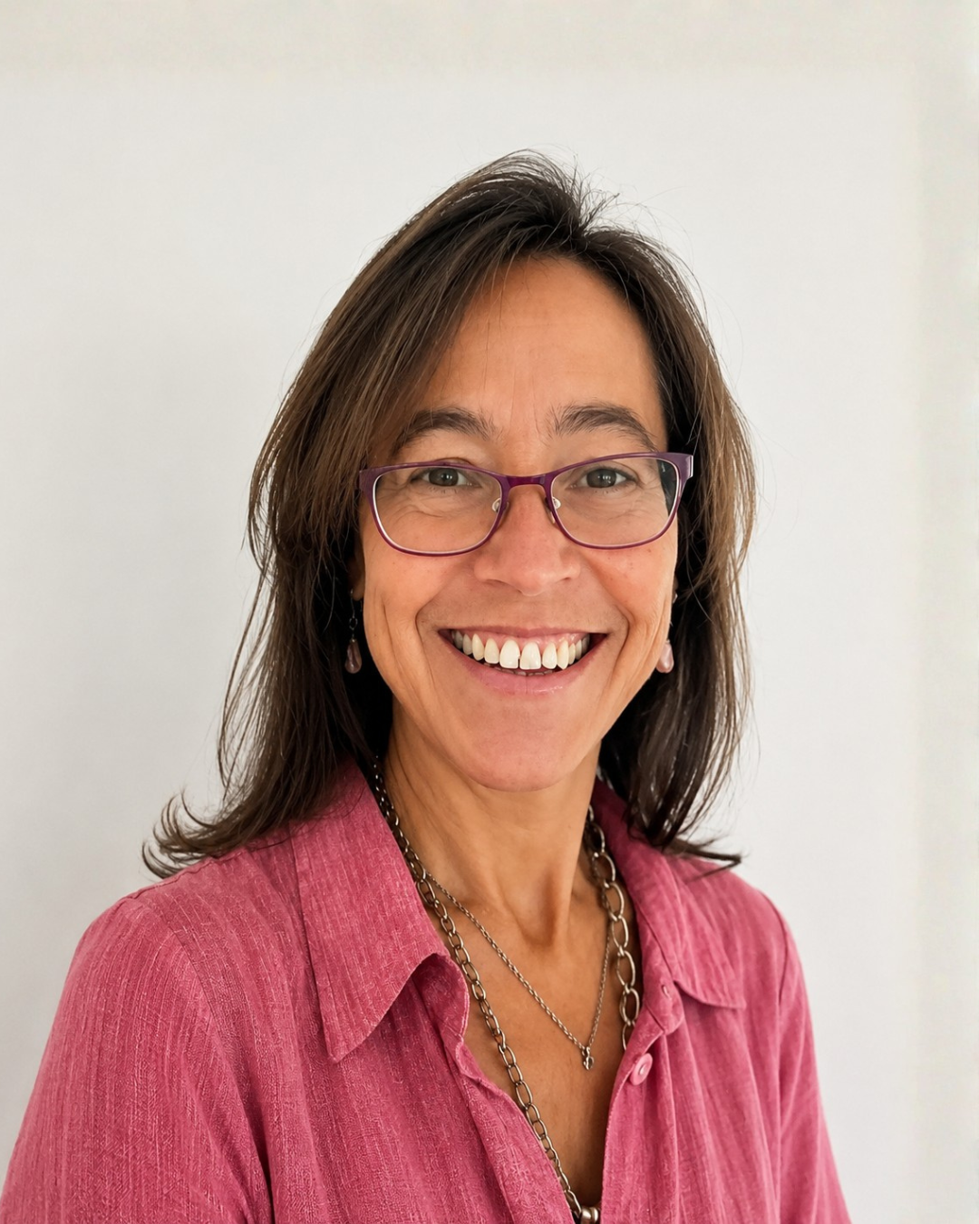 Retrato de una mujer sonriendo, con gafas moradas, ropa rosa y cabello castaño oscuro, frente a un fondo blanco.