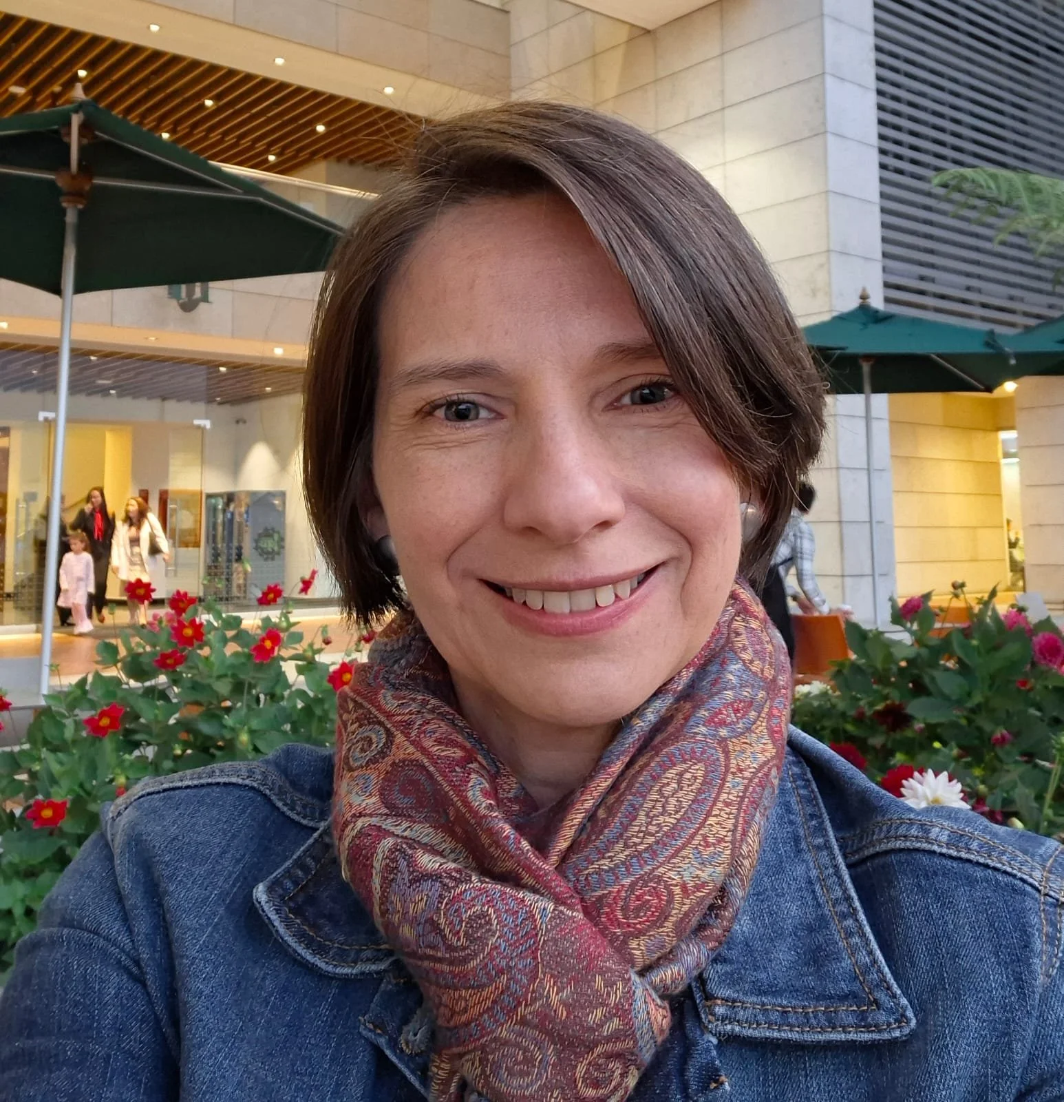 Retrato de una mujer sonriendo en un espacio interior con plantas y personas al fondo.