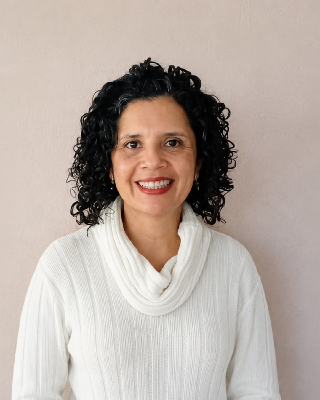 Retrato de una mujer con cabello rizado negro, sonriendo, usando suéter blanco, frente a una pared beige.