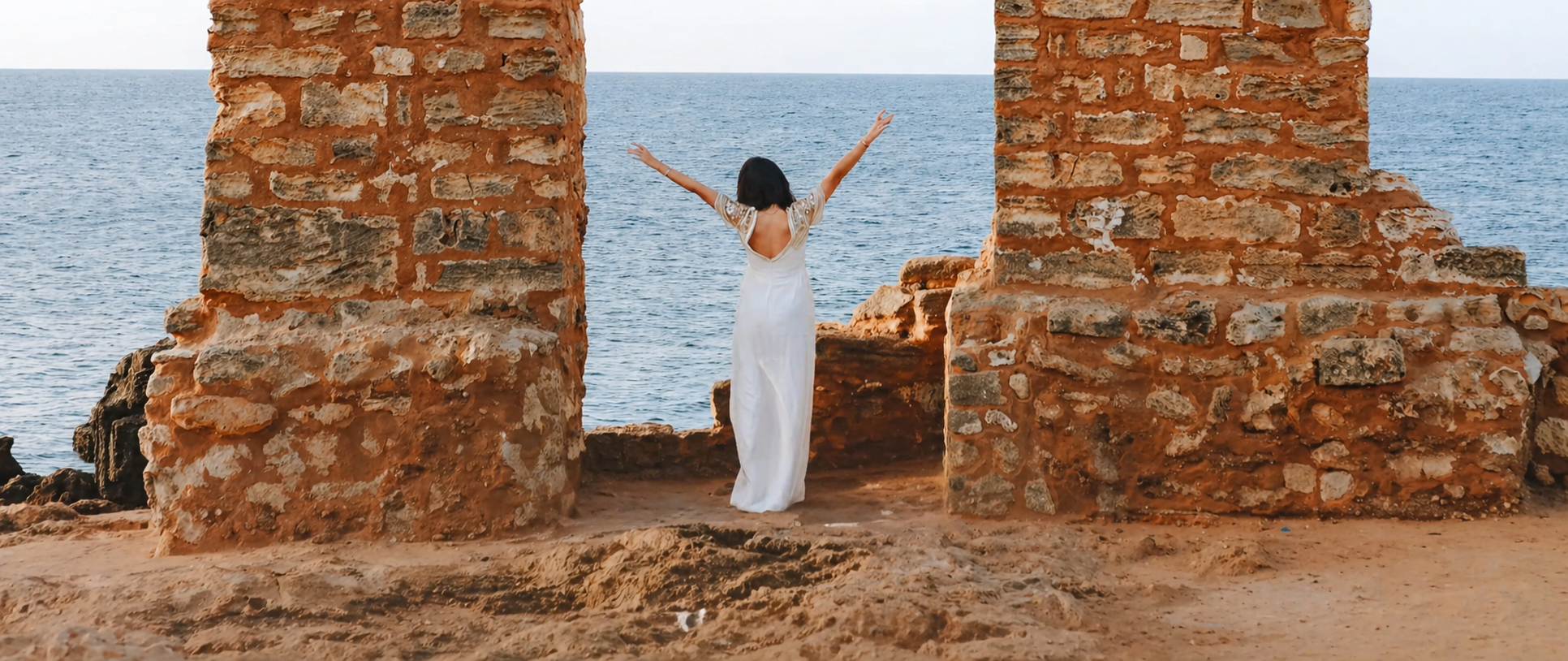 Mujer en vestido blanco, con los brazos levantados, de espaldas, entre ruinas de piedra junto al mar.