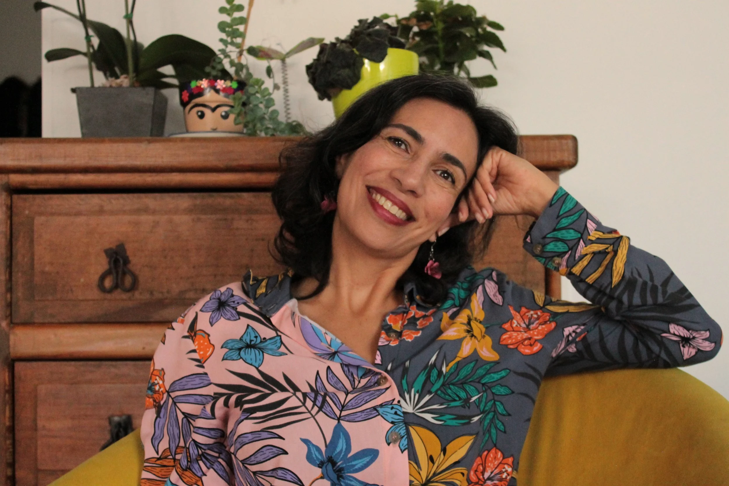 Mujer sonriendo, con cabello rizado negro y vestida con blusa de flores de diferentes colores, apoyada en su mano en una sala con fondo de pared blanca, una cómoda de madera y plantas en macetas.