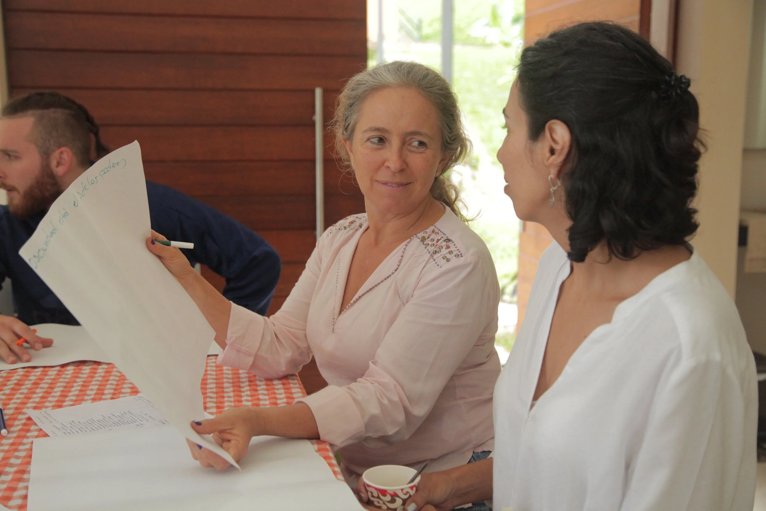 Dos mujeres conversando en una reunión, una de ellas está sosteniendo una hoja grande con escribiendo y la otra con una taza en la mano, en un ambiente interior con madera y luz natural.
