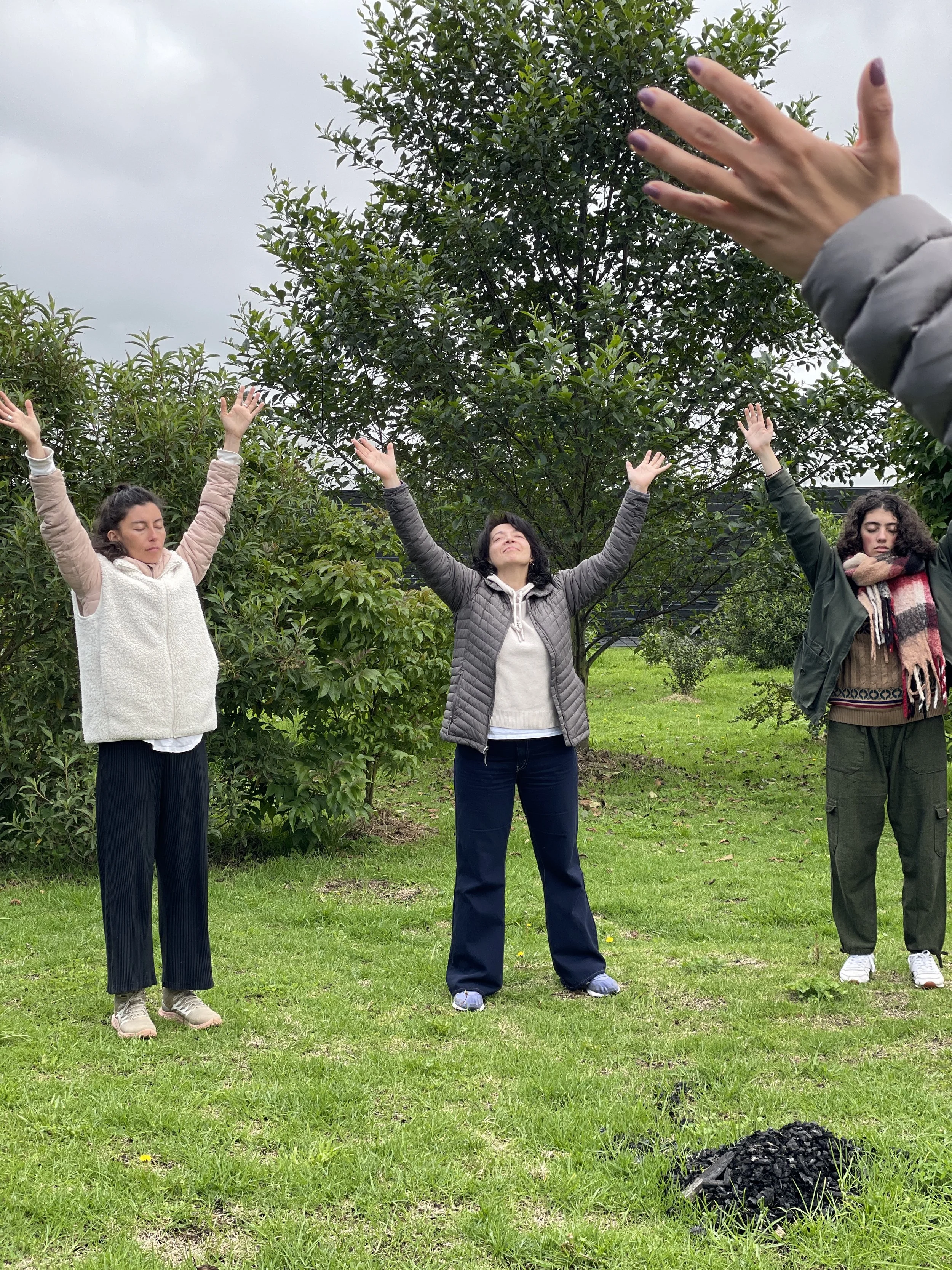 Tres mujeres de espaldas con los brazos levantados, en un prado con árboles y cielo nublado, con una pila de carbón quemado en el suelo.