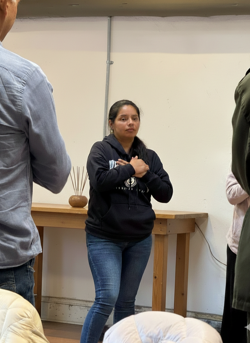 Mujer de pie con manos cruzadas en el pecho, en un ambiente interior, rodeada de otras personas, en un espacio sencillo con mesa de madera y decoración minimalista.