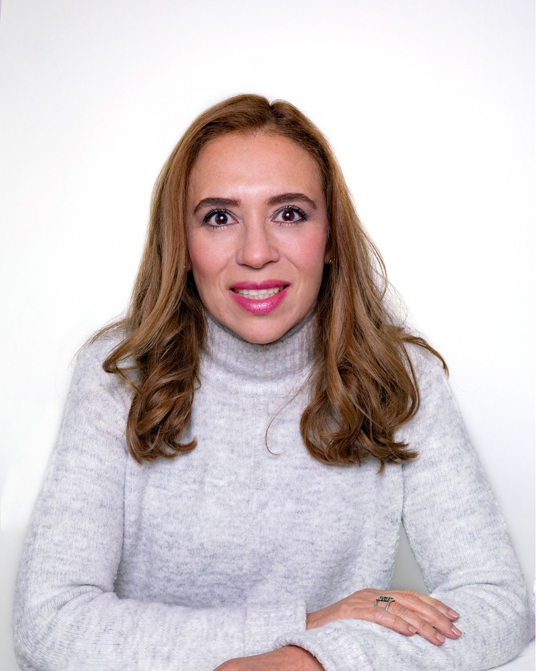 Retrato de una mujer con cabello castaño claro, cejas definidas, ojos grandes y sonrisa, usando un suéter gris claro, posando frente a un fondo blanco.