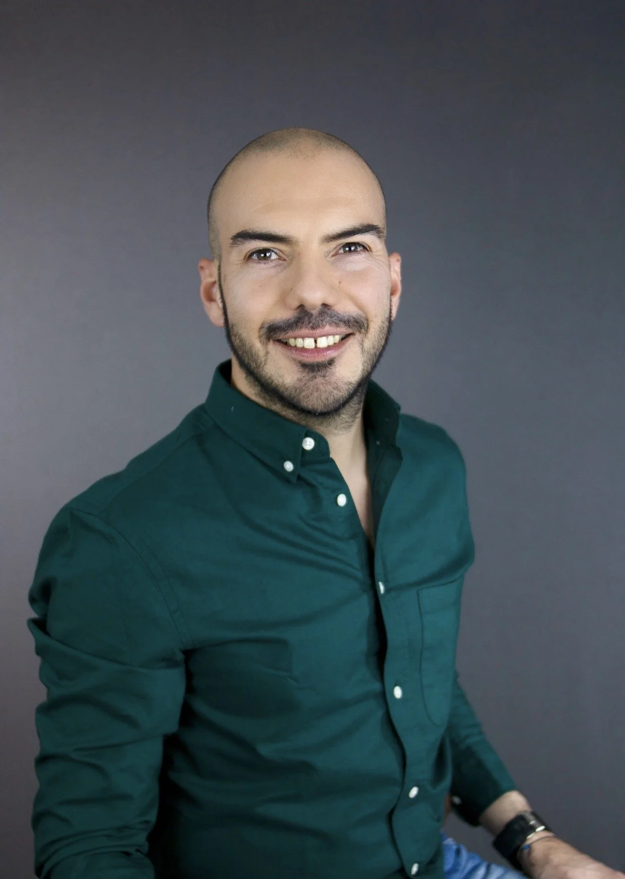 Hombre de barba y cabeza rapada sonriendo, vestido con una camisa verde oscuro, sentado contra fondo gris.