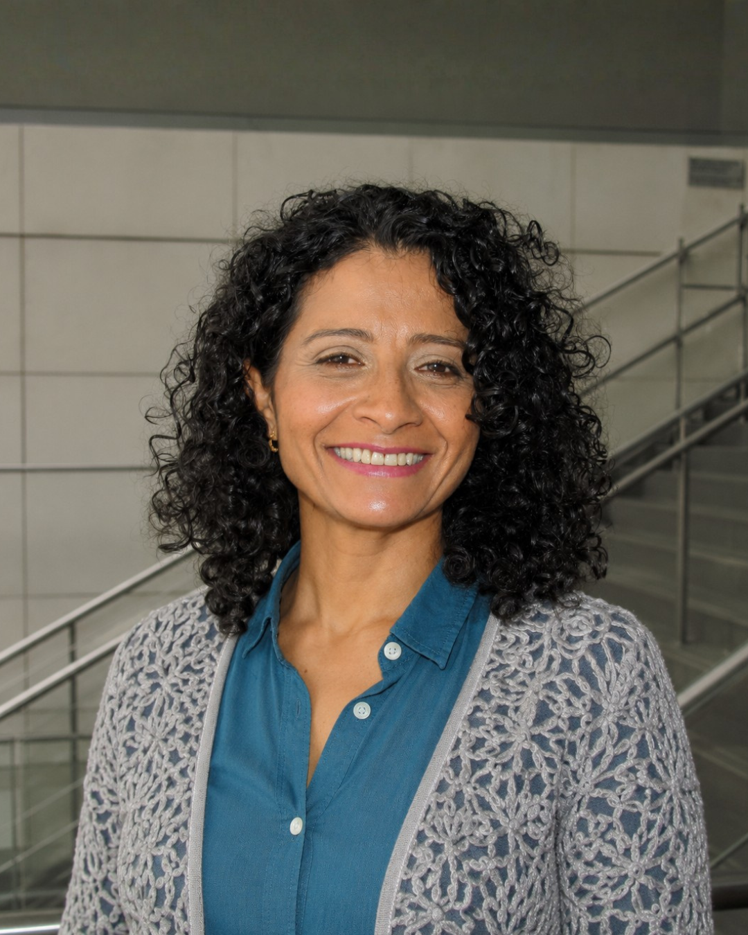 Retrato de una mujer con cabello rizado, sonrisa, vestida con camisa azul y suéter gris, en un espacio con escaleras metálicas y paredes de baldosas.