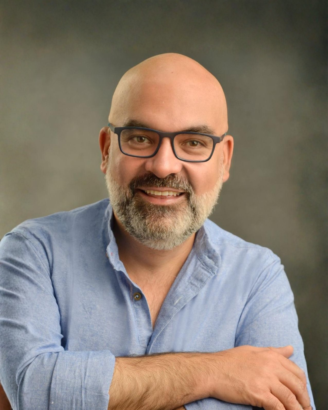 Hombre calvo con barba, gafas y camisa de mezclilla, sonriente, en retrato profesional.
