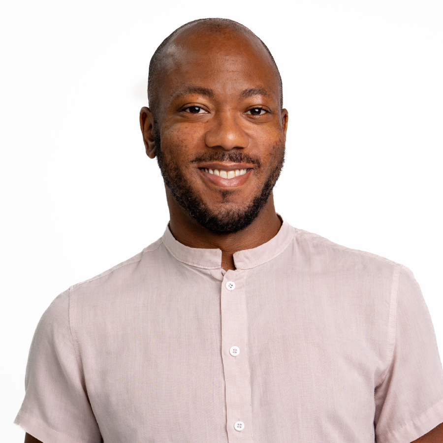 A smiling man with a beard and short hair wearing a light pink button-up shirt standing against a white background.