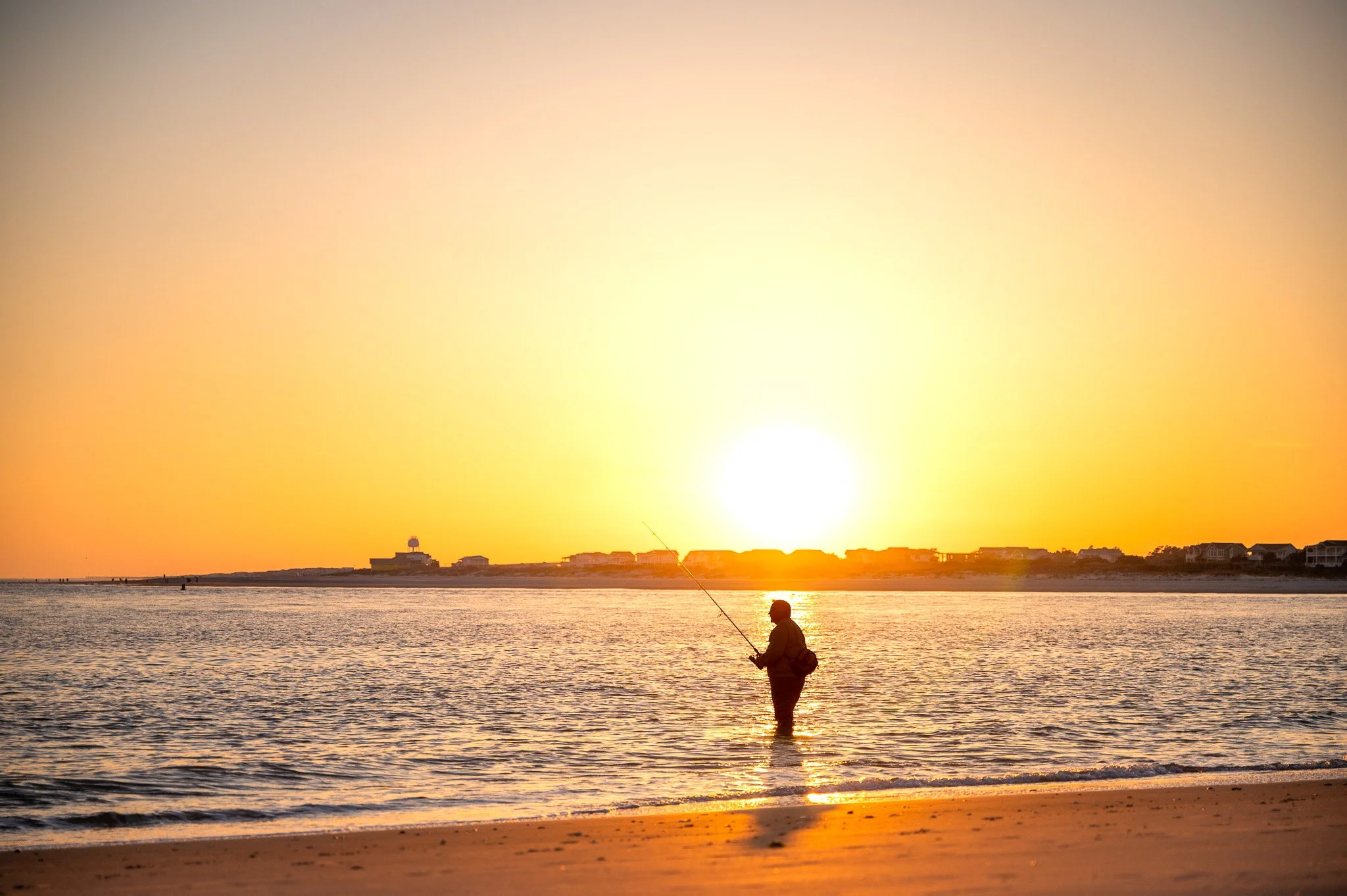 Angler fishing in shallow water at The Point on Oak Island, North Carolina, during a colorful sunset over the Intracoastal inlet.