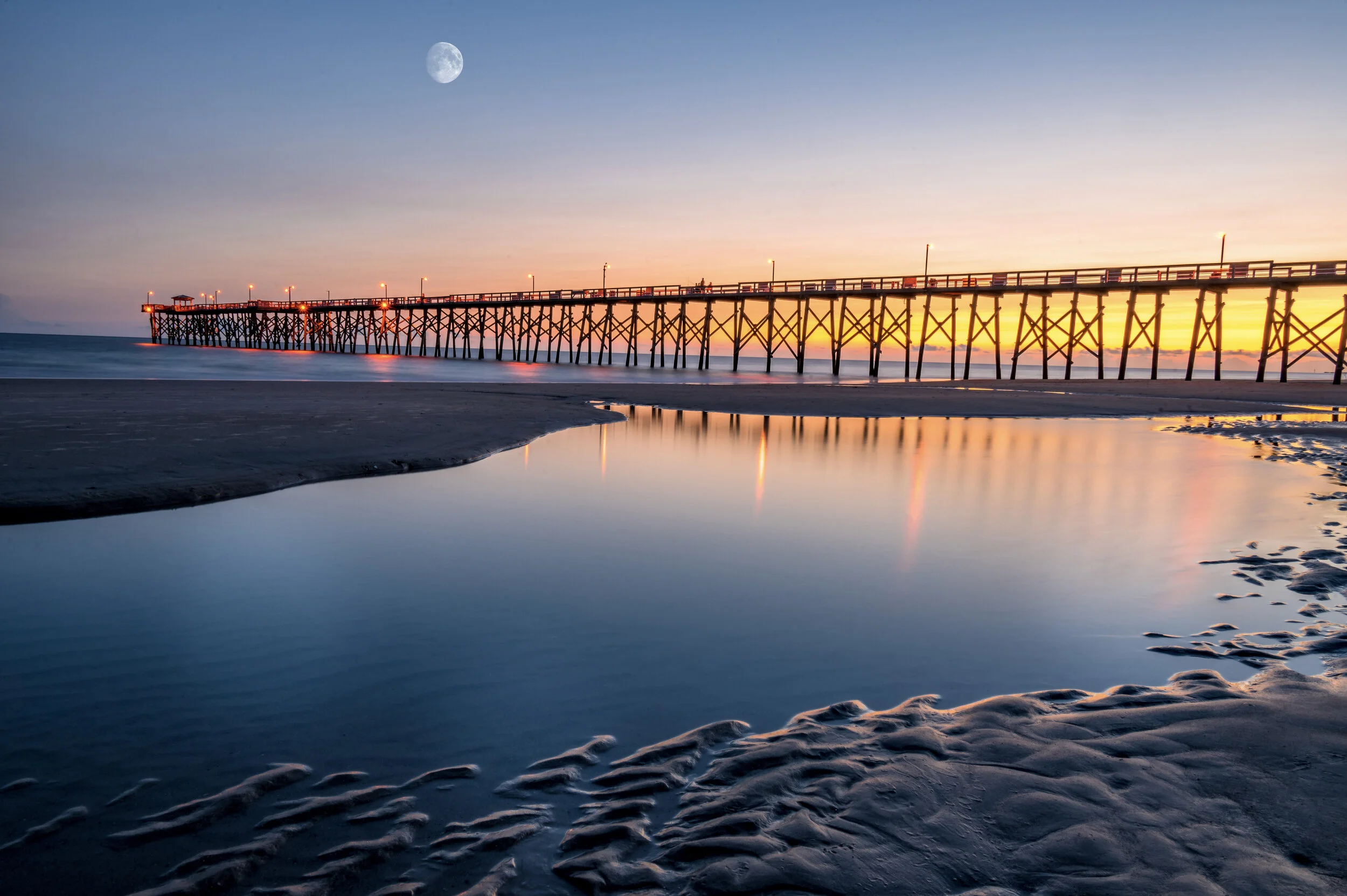 Oak Island Pier illuminated at dawn with reflections in tidal pools and the moon visible above the Atlantic Ocean in North Carolina.