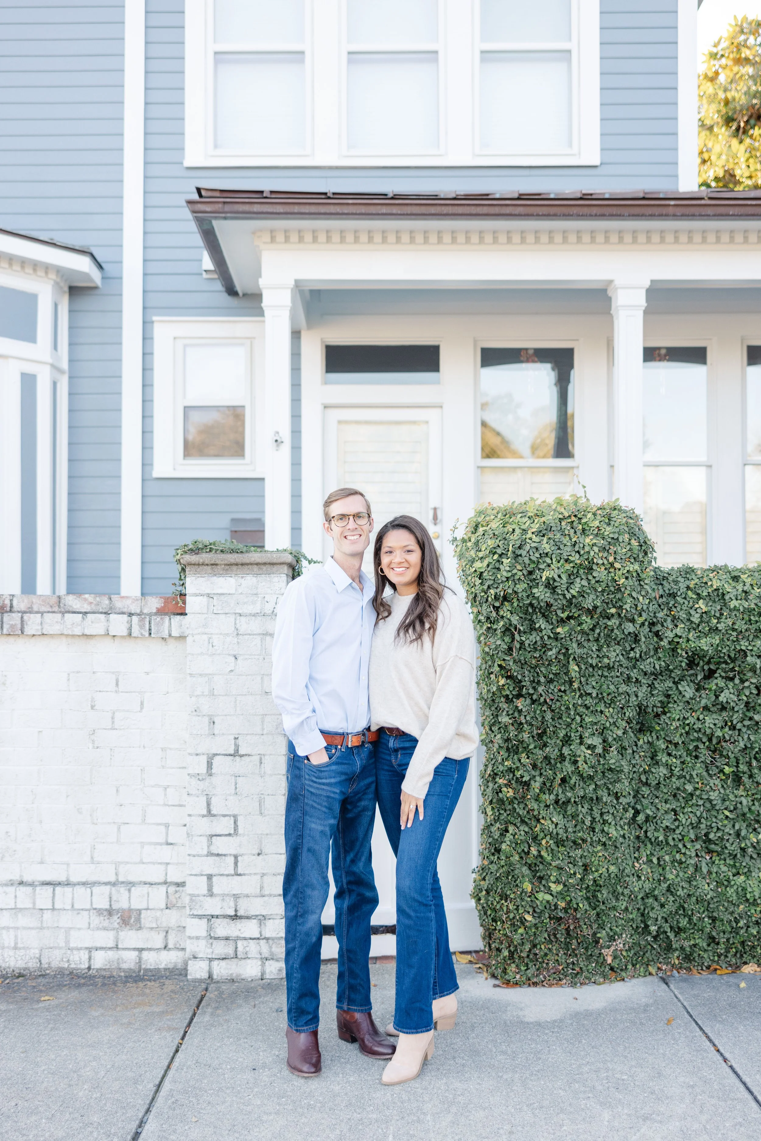 A smiling couple stands in front of a blue house with bushes and a short white brick wall on Downtown Southport, NC
