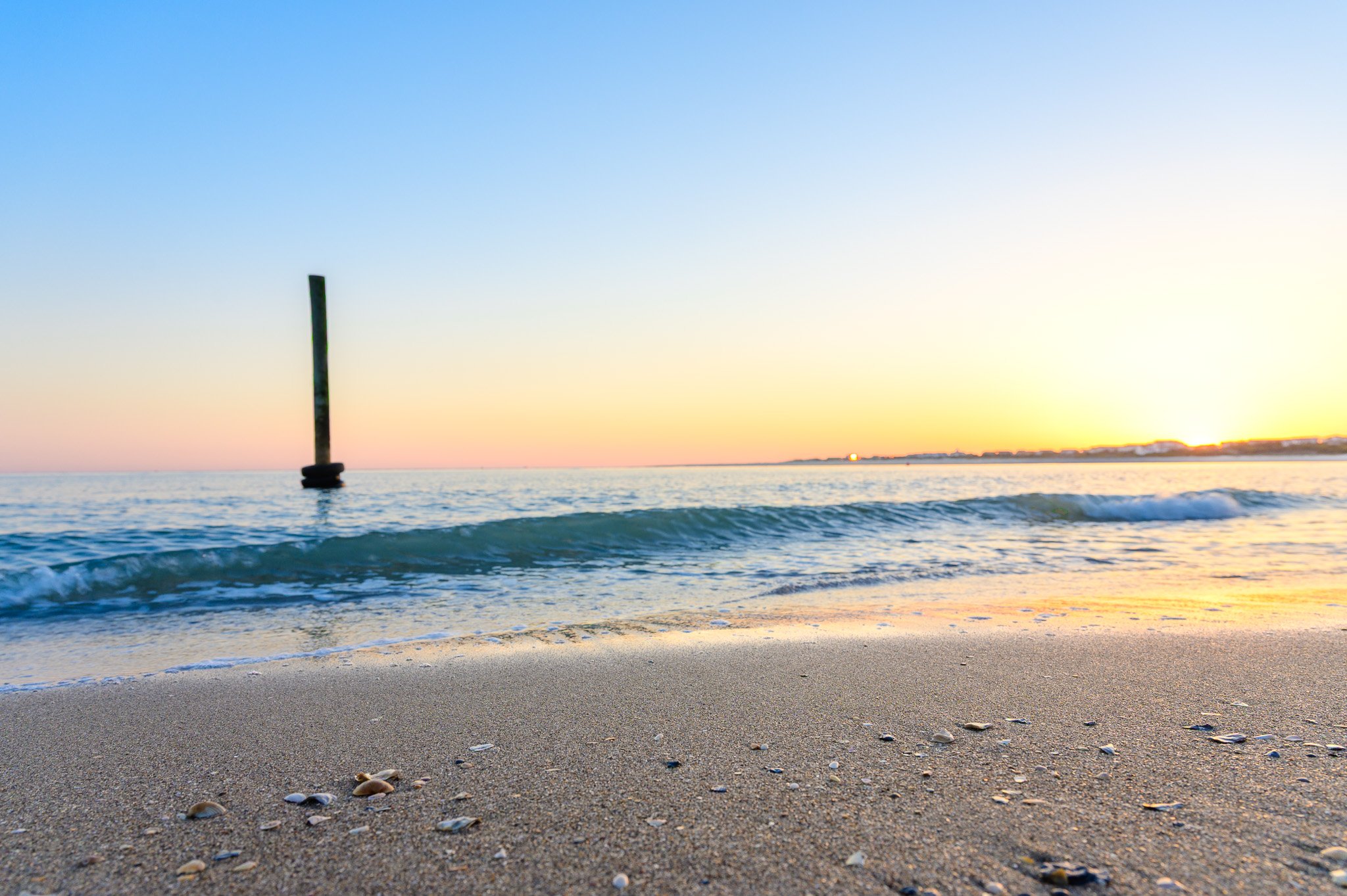 Seashells scattered along the shoreline at The Point on Oak Island, North Carolina, during a colorful sunset over the water.