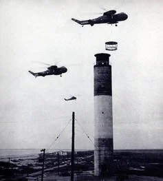 Historic photograph showing the Oak Island Lighthouse during construction in 1958 as helicopters lower the lantern room onto the top of the tower.