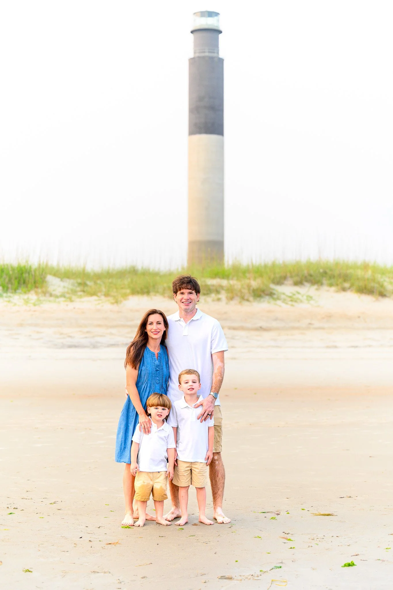 Family posing for beach portraits at Caswell Beach with the Oak Island Lighthouse visible in the background.