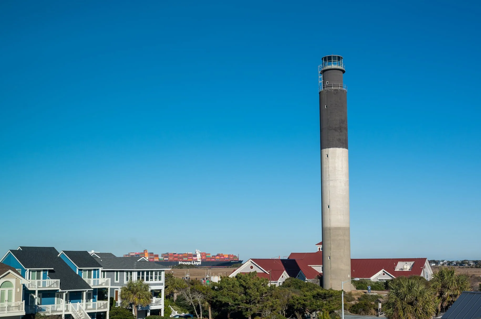 Oak Island Lighthouse towering above Caswell Beach with a container ship traveling through the Cape Fear River shipping channel in the background.